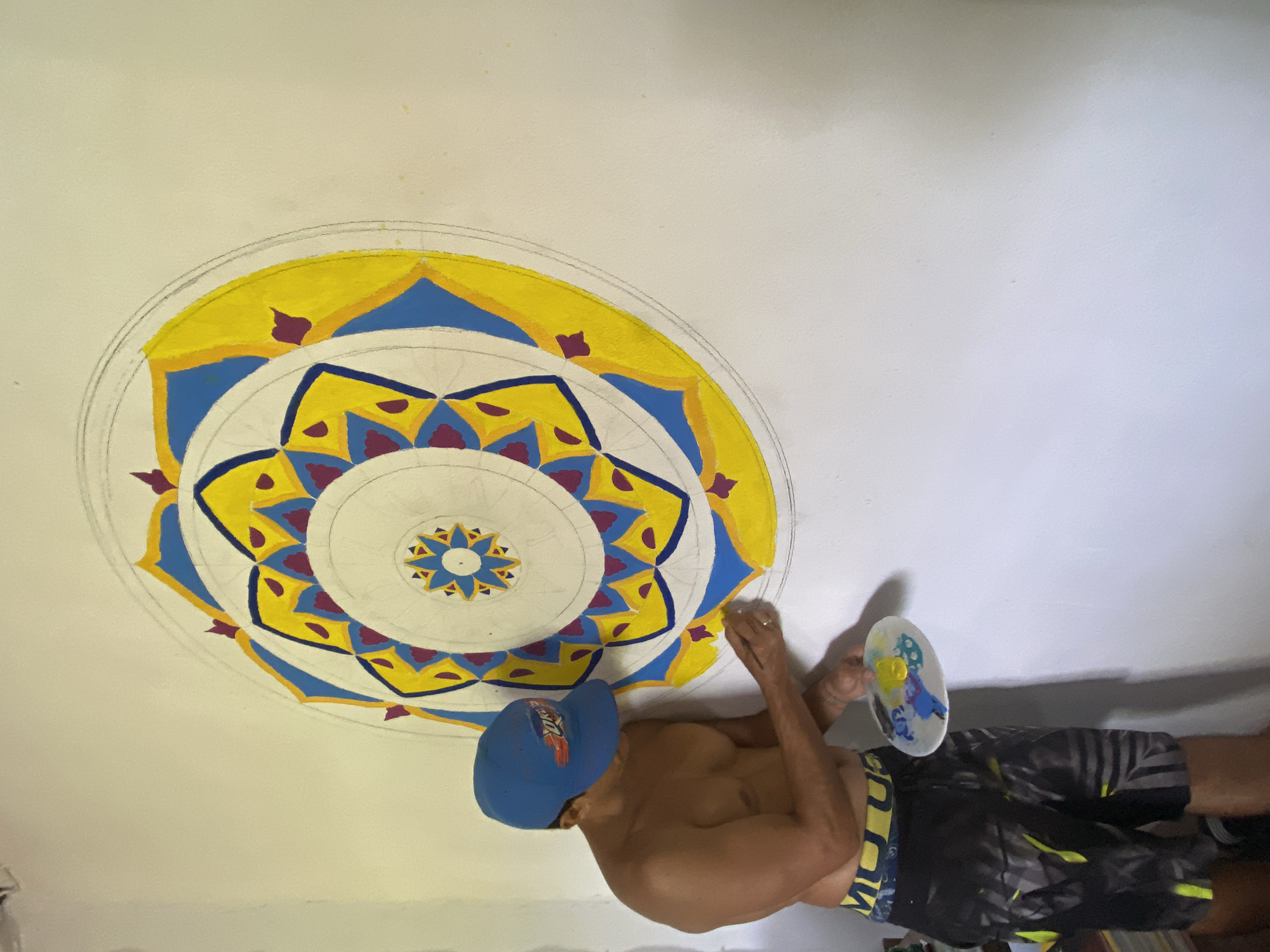 A shirtless man in a blue baseball cap and black shorts with yellow accents is painting a mandala on a white ceiling. He is holding a paint palette in his left hand and a paintbrush in his right. The mandala is composed of concentric circles and geometric shapes, primarily in yellow, blue, and maroon. The setting appears to be indoors. There is no indication of location or information to support location in Los Velásquez, Venezuela.