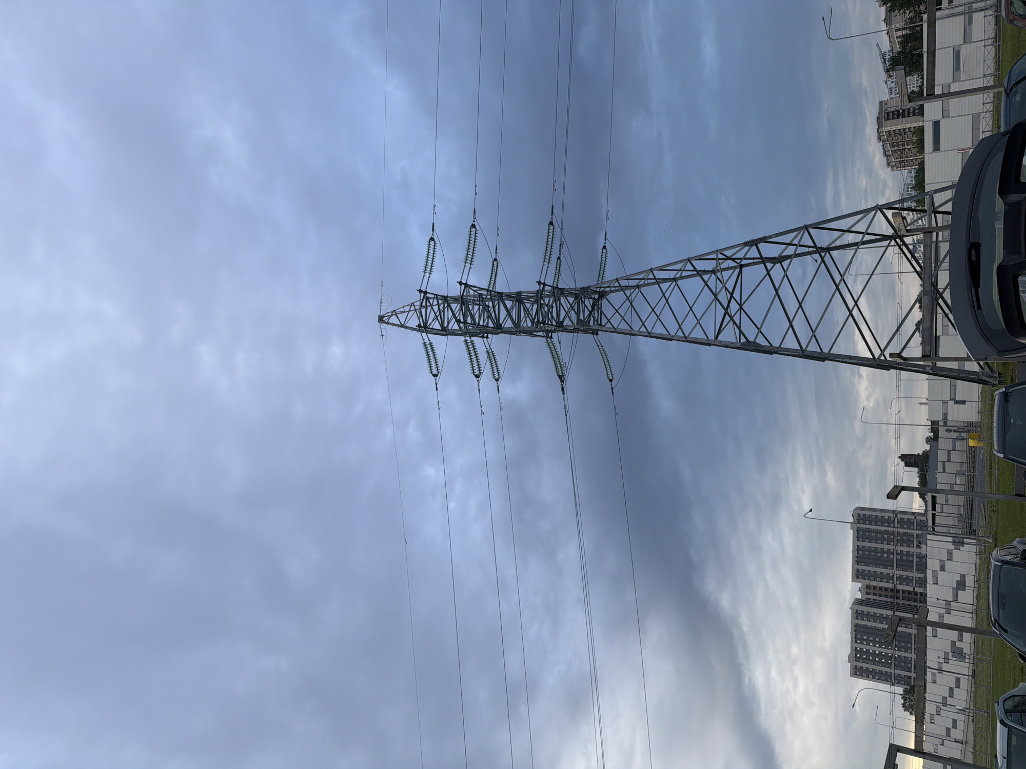 An outdoor, low-angle, full shot captures a tall, metal electricity pylon against a cloudy, gray sky. The pylon is centrally located and its lattice structure is clearly visible, supporting multiple power lines that stretch horizontally across the frame. Below the pylon, in the background, are several apartment buildings, a road, and parked cars. The overall scene depicts an urban or suburban environment with infrastructure for power transmission.