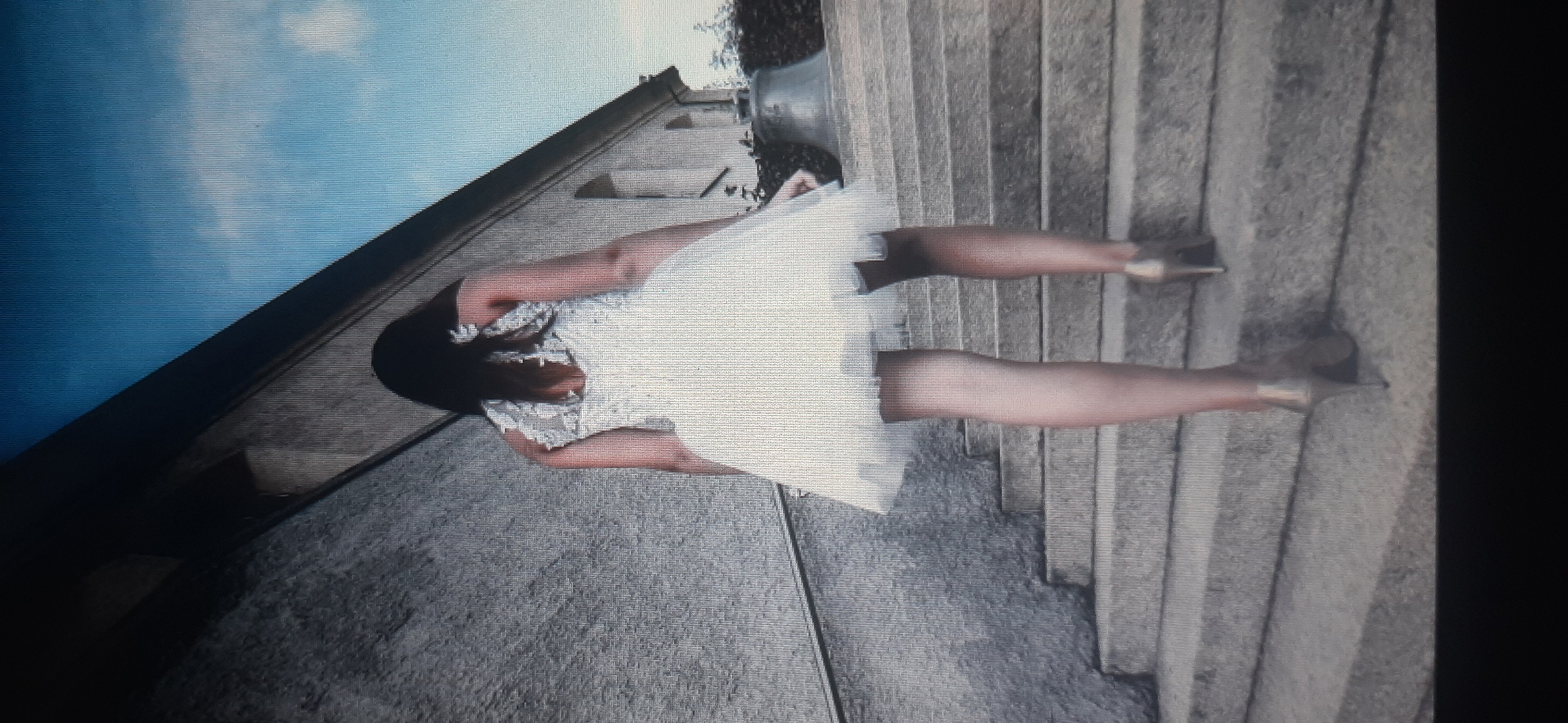 The image shows a woman from behind, wearing a short, white, puffy dress and high-heeled shoes, walking up a set of stone stairs. She has long, dark hair. The stairs lead to a building with a stone facade, and a bell is visible to the right of the stairs. The sky above is blue with some clouds. The location is not identifiable as Yessentukskaya, Russia.