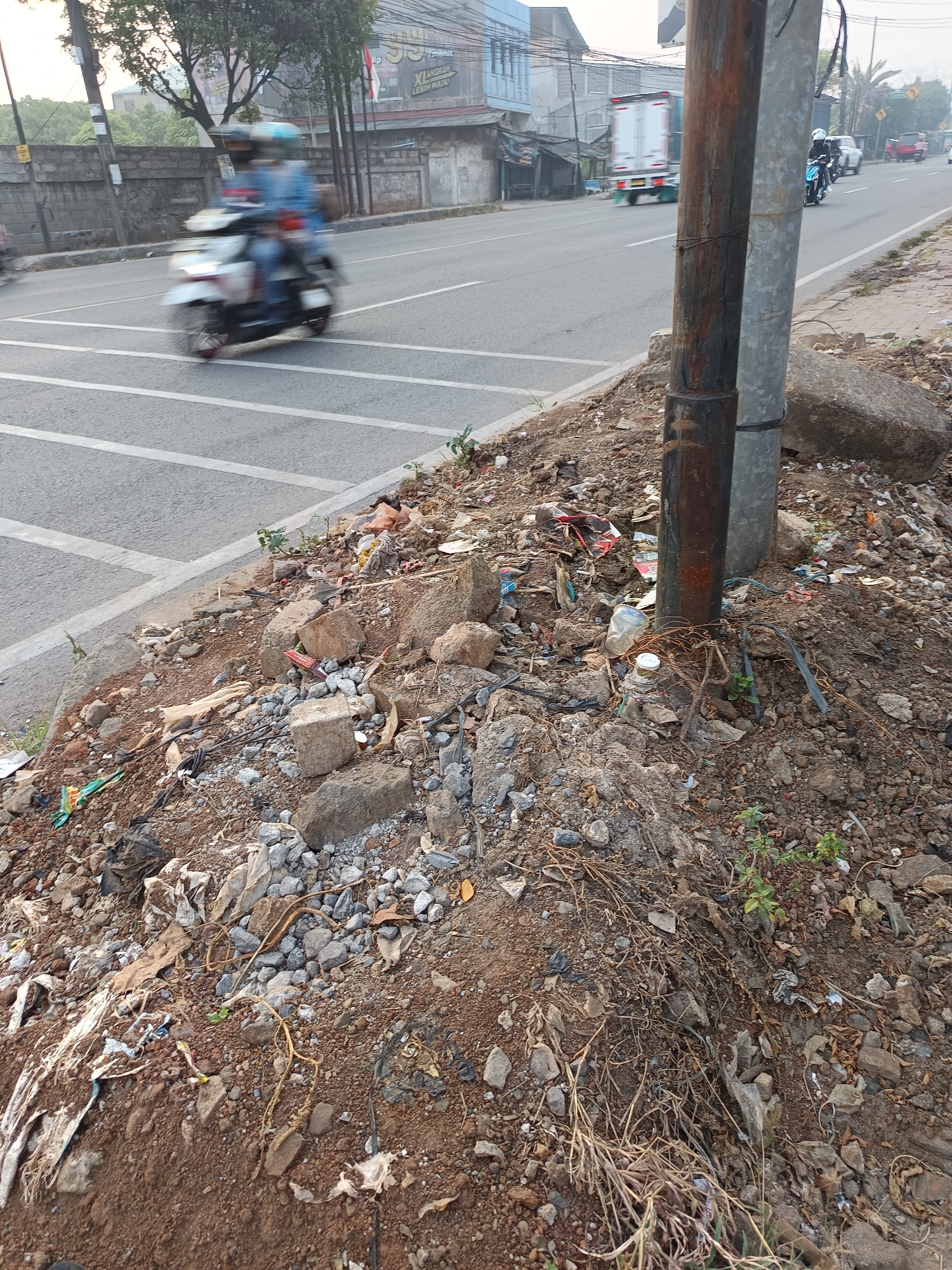 The image depicts a roadside area in Talaga Sari, Indonesia. In the foreground, the ground is covered with dirt, rocks, and a considerable amount of debris, including plastic bags, wrappers, and broken pieces of concrete. Two utility poles, one appearing rusted and the other metallic, stand prominently on the right side of the frame, partially obscured by the debris. A large boulder is visible behind the poles. In the background, a paved road runs parallel to the debris-laden roadside. Traffic is present, with a white truck in the middle distance and a white motorcycle with two riders blurred by motion in the left lane. Further down the road, more vehicles, including what appears to be a red pickup truck, are visible. Buildings and trees line the far side of the road, suggesting an urban or semi-urban environment. The lighting suggests it is daytime, possibly early morning or late afternoon due to the soft light.