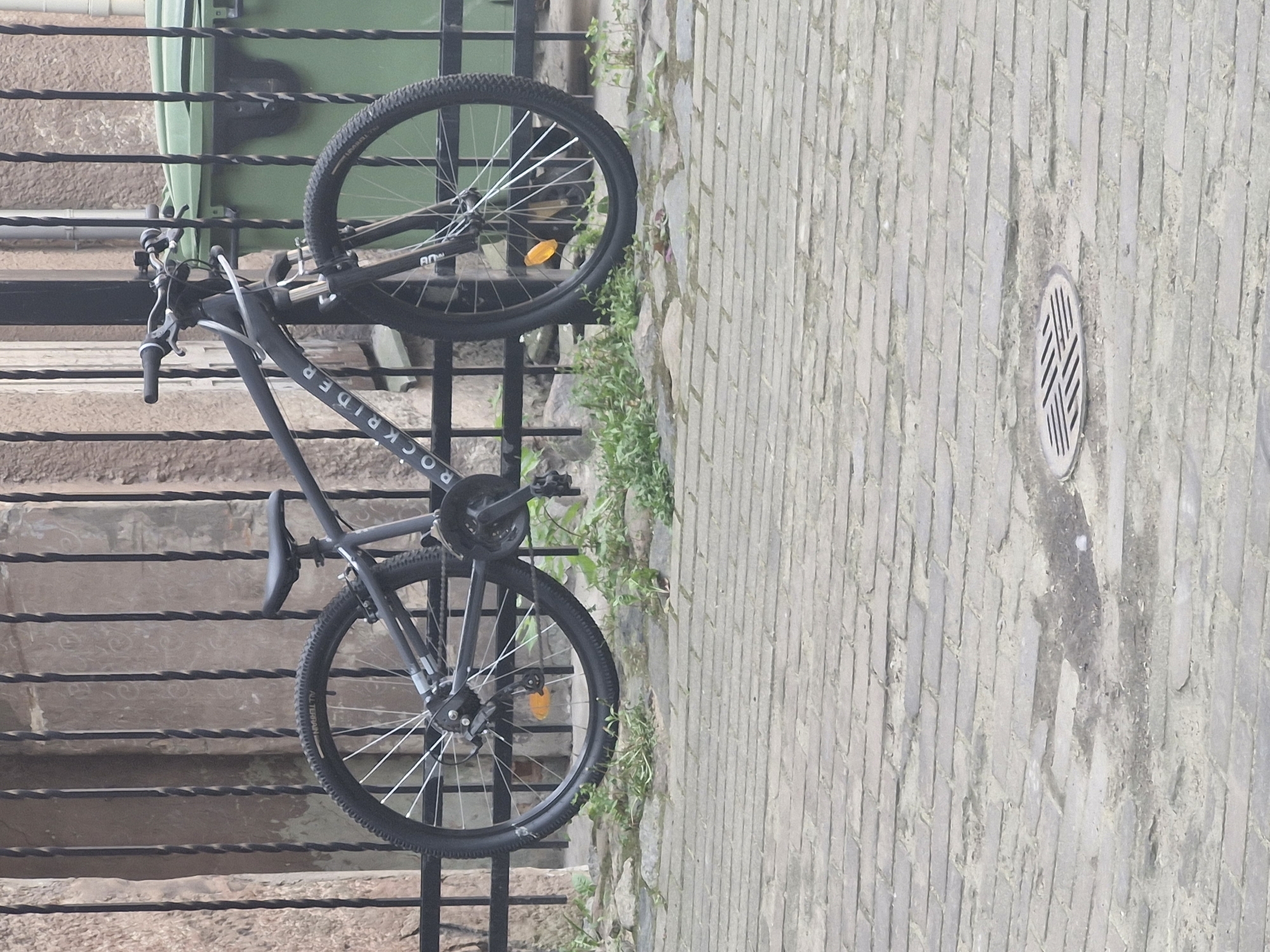 The image displays a dark grey bicycle positioned against a black metal fence or railing. The fence, which features twisted horizontal bars, is seen vertically in the frame. Behind the fence, a series of light brown concrete steps descends downwards. To the right of the bicycle and fence, a light grey paved surface extends vertically, resembling a wall, with rectangular blocks laid in a diagonal pattern. Small green plants grow sparsely along the base of this paved surface. A circular metal grate with a diagonal cross-hatch pattern is embedded in the paved surface.
The bicycle has "ROCKRIDER" printed in white letters on its frame. Both front and rear wheels are visible, and the tires bear the text "ALL TERRAIN." An orange reflector is attached to the spokes of the front wheel. Other bicycle components such as handlebars, seat, pedals, and chain are also clearly visible. The lighting suggests it is daytime, with soft illumination and no distinct shadows. There are no people present in the scene.