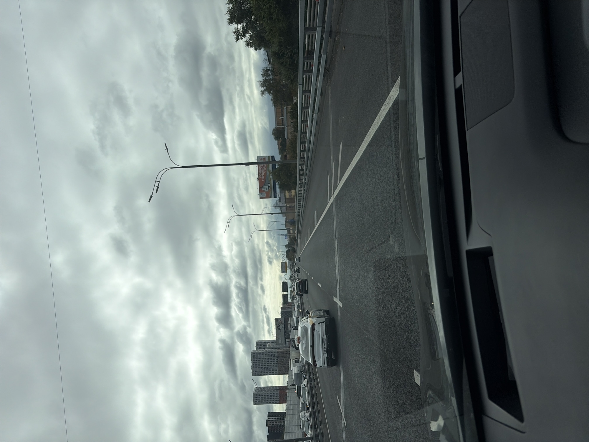 The image depicts a view from inside a vehicle on a multi-lane highway in Moscow, Russia. The sky is overcast with heavy, gray clouds. On the left side of the highway, there are several tall, modern buildings, some of which appear to be residential complexes. To the right of the highway, there is a metal guardrail and beyond that, trees and some low-lying structures. A billboard with Russian text is visible in the distance. The highway itself is paved and has white lane markings. Several vehicles are on the road, including a white Kia sedan with a yellow taxi sign on its roof, which is in the foreground and appears to be the vehicle the photo is taken from. The overall impression is of a typical urban highway scene under cloudy weather.
