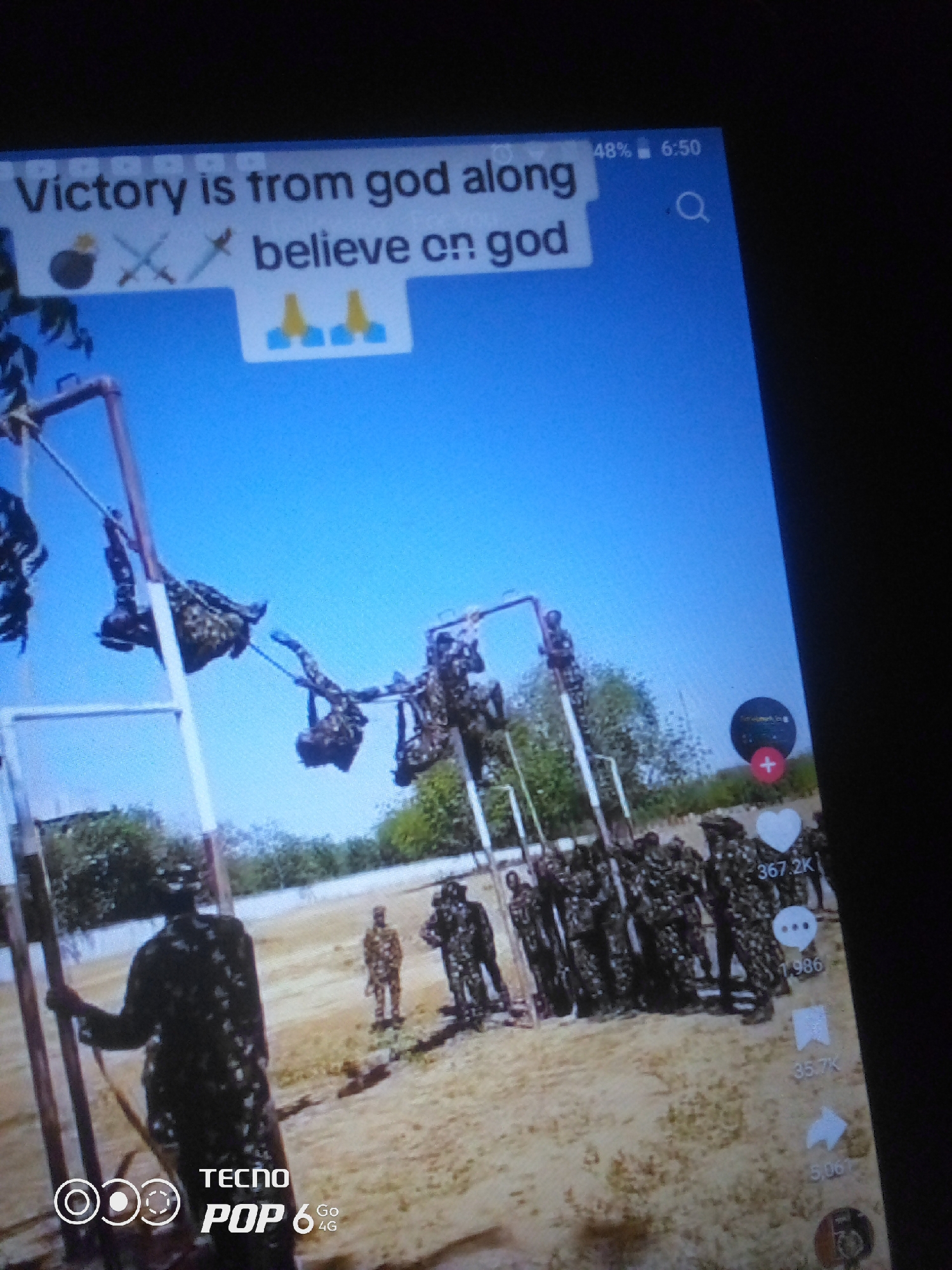 The image displays a mobile phone screen with a video playing. The video shows a group of soldiers in camouflage uniforms engaged in a training exercise. Some soldiers are seen suspended from a structure that appears to be an obstacle course, possibly performing some form of agility or strength training. Other soldiers are standing in formation on a sandy ground. The background consists of a clear blue sky and some sparse vegetation. Text overlays on the screen include a caption reading "Victory is from god along believe on god" with emojis and a search icon. Social media interaction icons and numbers, such as a heart with "367.2K" and a speech bubble with "1.986", are also visible on the right side of the screen. The bottom of the screen shows the TECNO POP 6 4G branding. The location of Howayi, Nigeria, is not discernible from the visual information presented in the image.