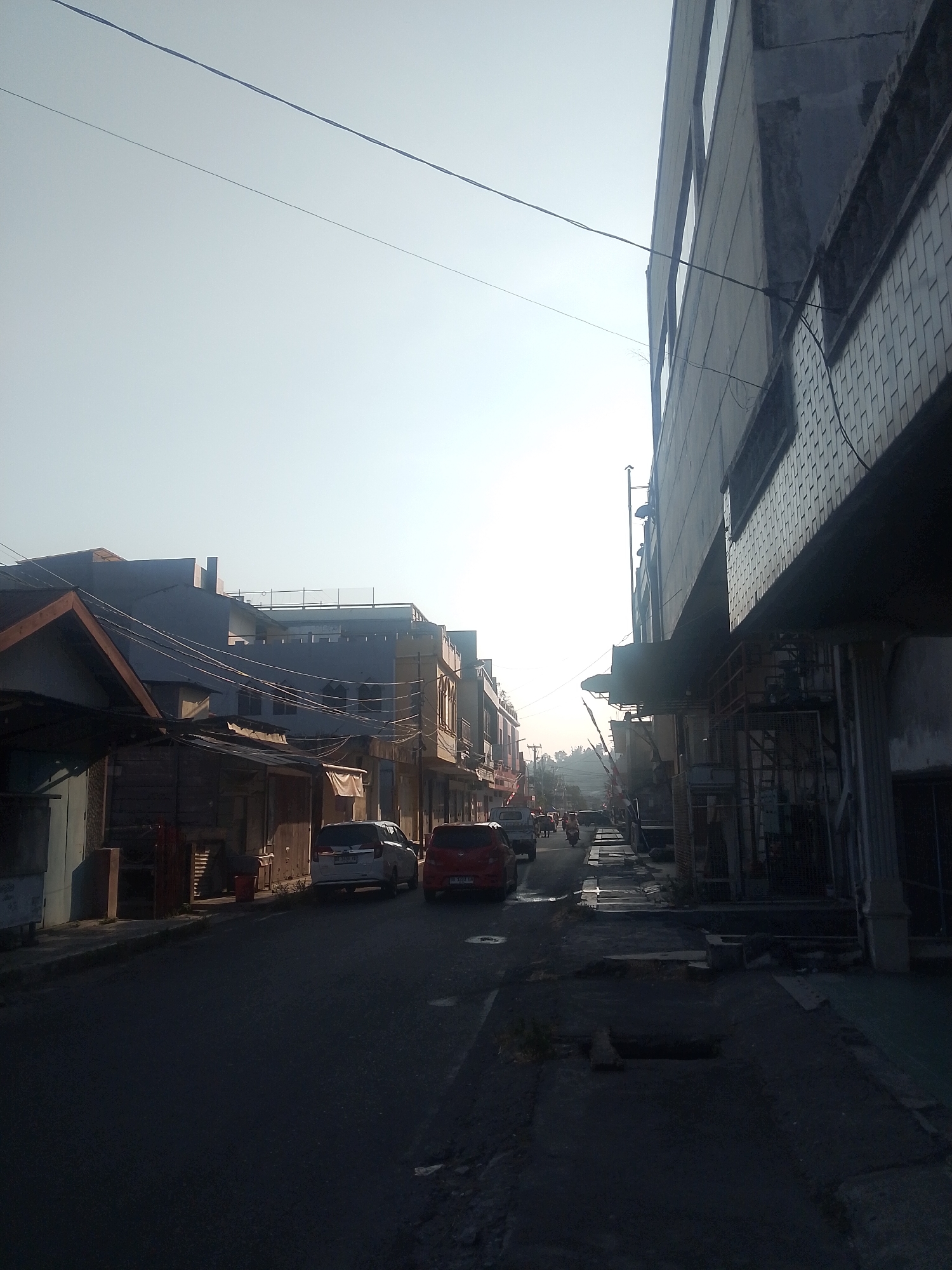 This is a low-angle, eye-level, full outdoor shot of a street in Uwuran Satu, Indonesia, at approximately 6:30 AM, as indicated by the direction and intensity of the sunlight. The sky is clear and bright, with a slight haze. The street is paved with asphalt and appears to be in moderate condition, with some patches of unevenness and debris.
On the left side of the street, there are several buildings. The closest building is a single-story structure with a wooden facade and a sloped roof. It appears to be a small shop or stall, with open doorways revealing cluttered interiors. Behind this structure, there are multi-story buildings with grey and white facades, featuring multiple windows and balconies. Power lines are strung across the street, connecting various poles and buildings.
On the right side of the street, a multi-story building dominates the foreground. Its facade is textured with small, rectangular tiles, and it has a series of large windows. The building's ground floor appears to be occupied by shops or businesses, with metal grates and awnings visible. Further down the street on the right, more buildings of varying heights and architectural styles can be seen.
There are several vehicles on the street. In the mid-ground, a white SUV and a red sedan are parked. Further down the street, a few motorcycles and cars are moving. The presence of traffic suggests the street is active.
The overall atmosphere of the scene is one of a typical urban environment in Indonesia waking up to a new day. The lighting suggests early morning, with long shadows cast by the buildings. The composition emphasizes the street and the architecture of the surrounding buildings, with the sky serving as a backdrop.