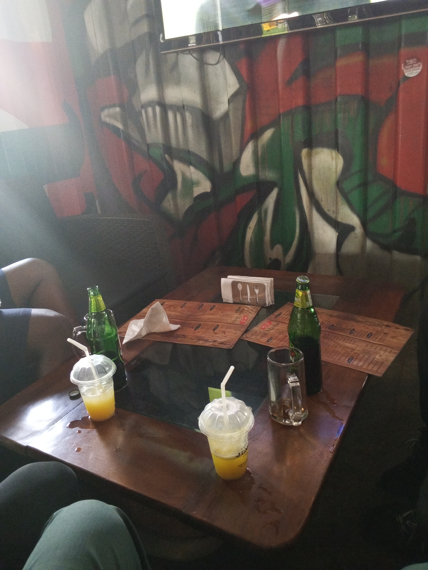 The image shows a wooden table in an indoor setting with graffiti on the walls. Two clear plastic cups filled with a yellow liquid are on the table, each with a white straw. Two green glass bottles, one with an attached glass handle and a clear glass mug. There are brown paper menus on the table. There are paper napkins in a holder and lying on the table. Parts of people's legs and clothing are visible around the table. The location is likely a bar or restaurant in Abuja, Nigeria.