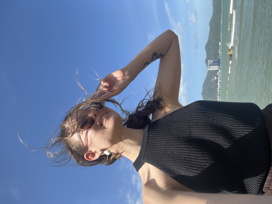 A female subject is depicted in a bright, outdoor coastal setting in Nha Trang, Vietnam. She occupies the foreground, positioned to the right of the frame's vertical axis, facing right with her head tilted slightly upward and a visible smile. Her light brown hair is actively blown upwards and to the left by the wind. She wears dark sunglasses with light-toned frames. A white, irregularly shaped earring adorns her left ear. Her attire consists of a black, knitted halter-neck top. Her right arm is bent at the elbow, with her hand raised near the side of her face, fingers touching the temple area. A dark, serpentine tattoo is visible on the inner bicep of her right arm. The midground features ocean water with moderate waves, some crashing against exposed rocks, creating white spray. The background reveals a distant mountainous coastline featuring various structures, including a prominent white high-rise building. The sky overhead is clear and blue.