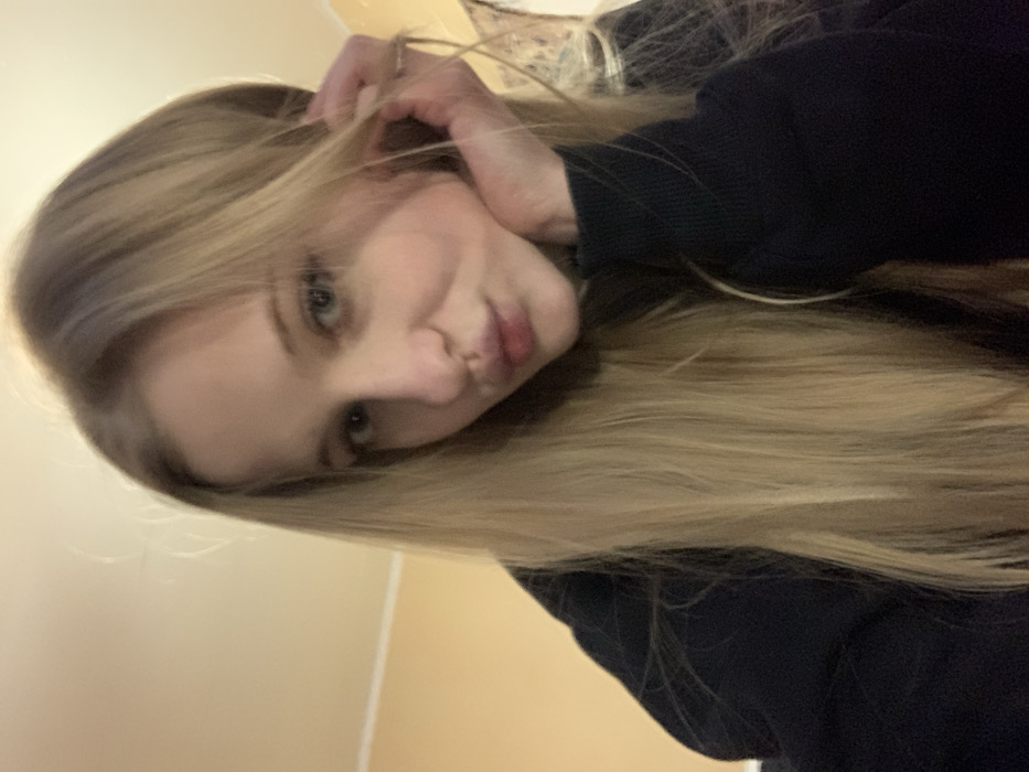 A blonde woman tilts her head, puckering her lips towards the camera. Her hand is up, fingers playing with her hair. She wears a dark-colored long-sleeved shirt. The background consists of a light-colored wall. The photograph is oriented with the woman's head tilted slightly to the right, making the image appear rotated.