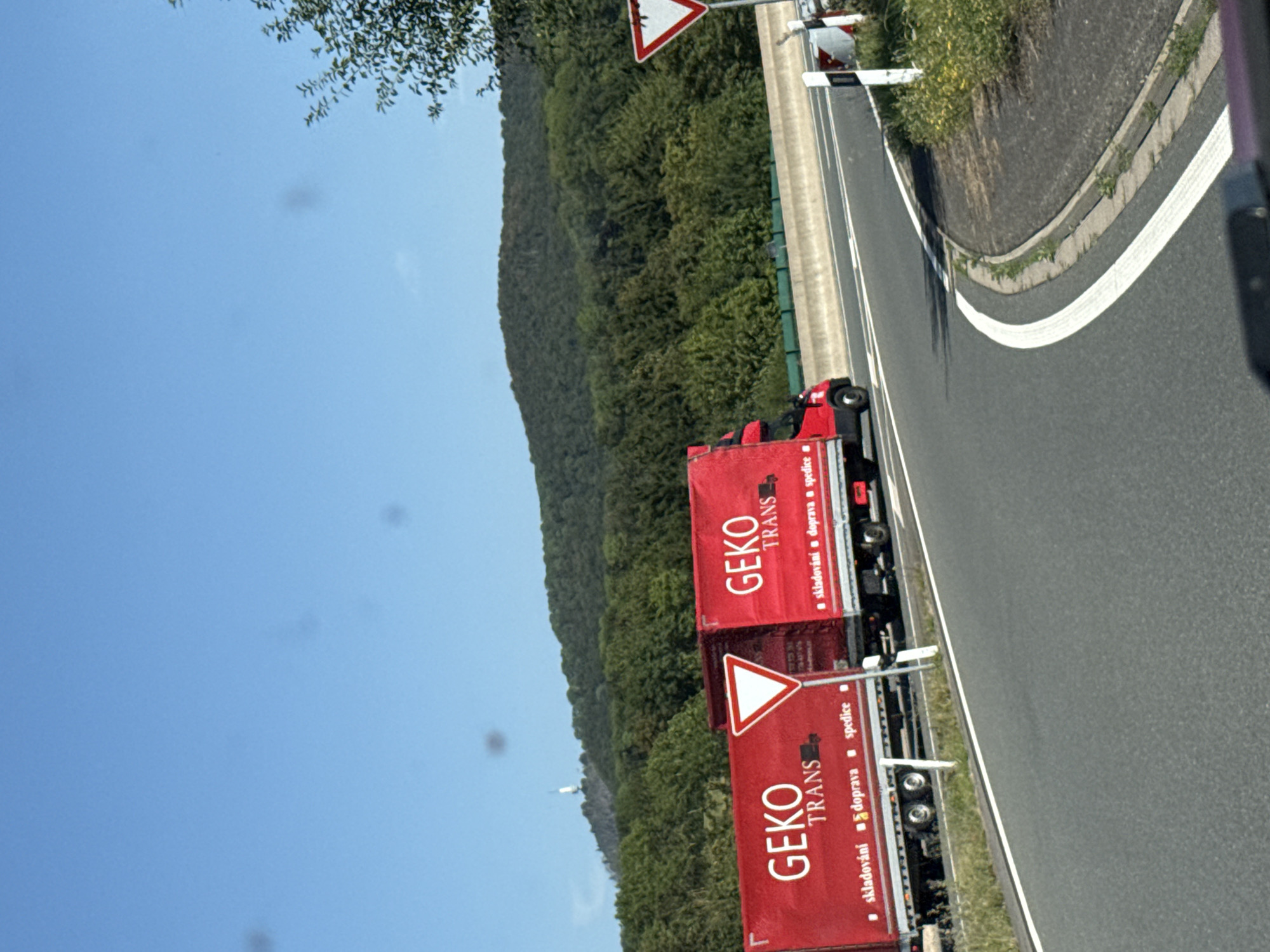 An outdoor, eye-level, medium shot captures a sunlit highway scene. The image is dominated by two bright red semi-trucks on the right side of the road, each with "GEKO TRANS" written in white lettering. The truck on the left has additional white text that includes "skladováni doprava spedice". A white triangular yield sign is positioned between the two trucks. The trucks are traveling on a highway with a concrete barrier to their right. In the background, a lush green forest covers a rolling hill under a clear blue sky. A white tower-like structure is visible on the hilltop. The foreground shows the asphalt of the highway and its white lane markings. This scene is likely located on a highway near Bad Lauterberg im Harz, Germany, given the context.