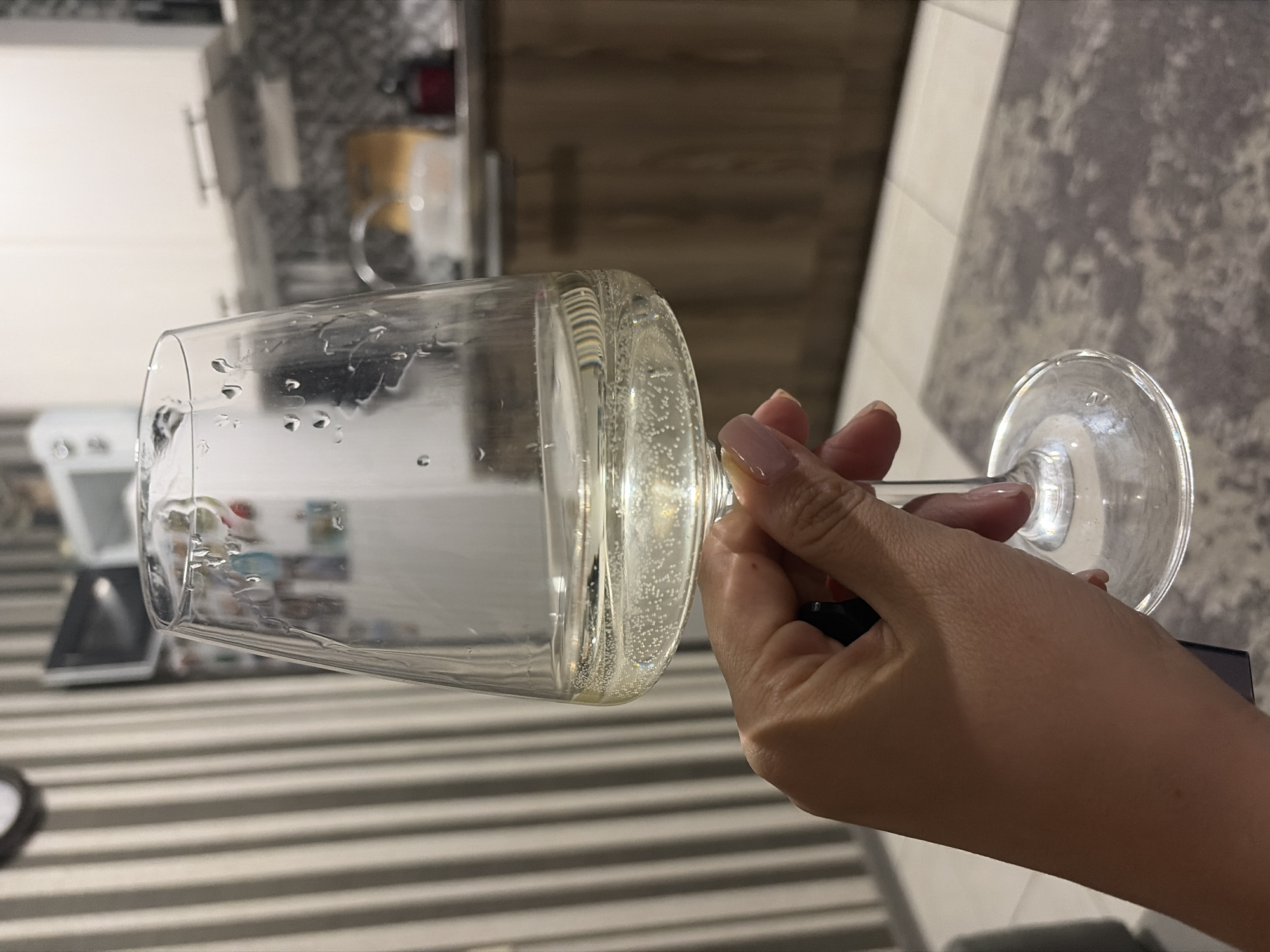 A woman's hand is holding a stemmed wine glass filled with a pale yellow, bubbly liquid. The glass is partially covered in condensation droplets. In the background, a kitchen is visible with light-colored cabinets, a dark countertop, and a tiled backsplash. A striped curtain hangs to the left of the frame, and a rug is on the floor. There is no indication of the image being located in Sochi, Russia.