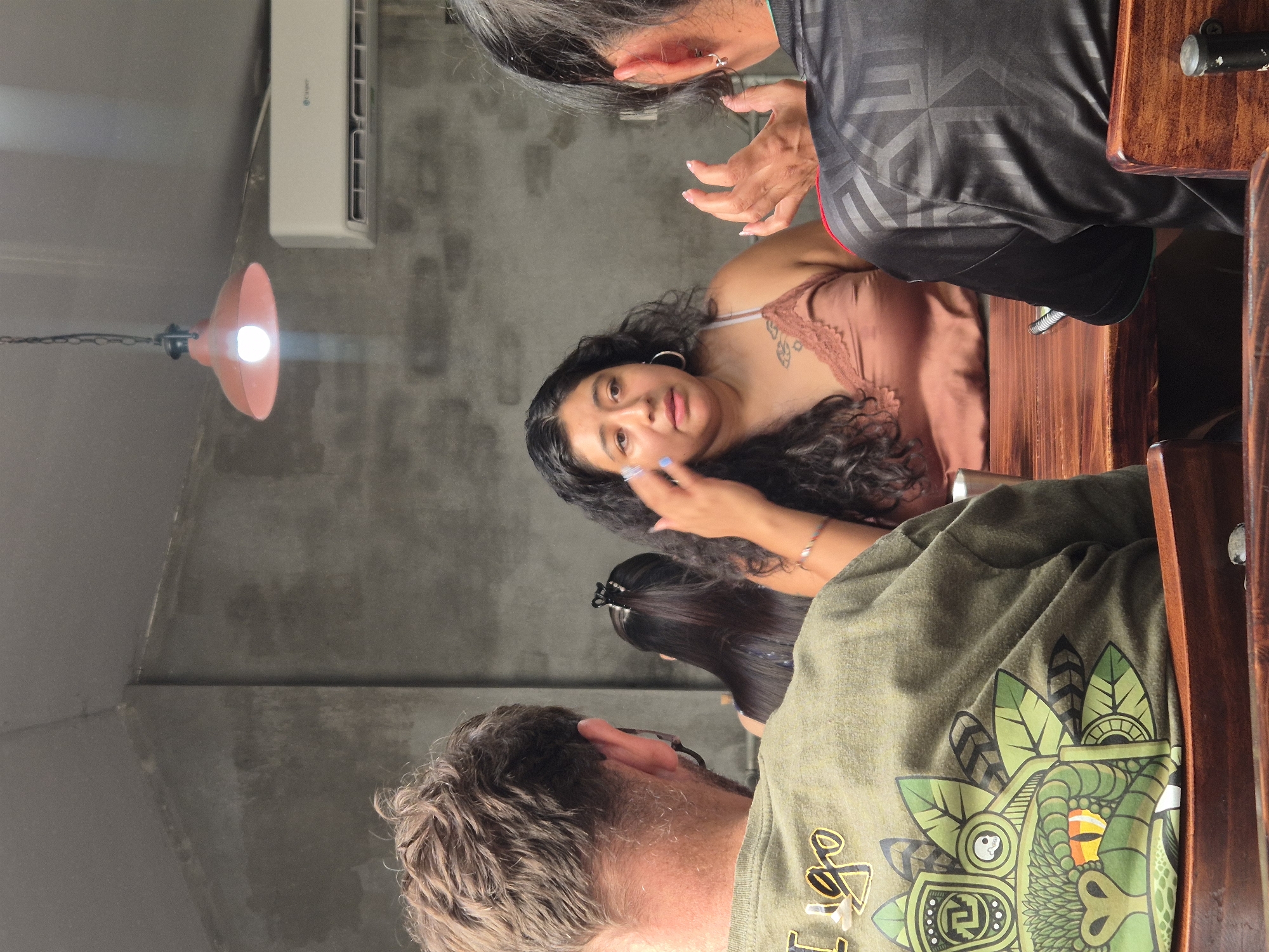 In Đà Nẵng, Vietnam, four individuals are seated at a wooden table. Two are seen from the back, one wearing a green t-shirt with a graphic of a stylized snake head and leaves, and the other in a black t-shirt with a geometric pattern. A woman with long, dark, curly hair is in the center, wearing a rust-colored camisole and touching her face. Another person with long dark hair is partially visible in the background. The environment appears to be an indoor dining area with a textured concrete wall and a hanging pendant light. An air conditioning unit is mounted on the wall. The focus is on the group engaged in conversation, with a slight blur affecting some areas of the image.