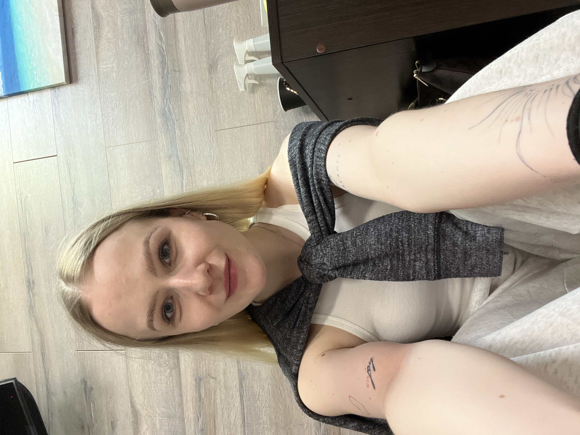 A fair-skinned female with blonde hair is taking a selfie. She is wearing a white tank top and a gray sweater that is tied around her neck. Tattoos are visible on her arms. A dark wooden cabinet is to her right, with cleaning spray bottles on the floor next to it. A partial painting is visible on the wall to her left, and the floor appears to be light wood laminate. No information in the image indicates this is in Khimki, Russia.
