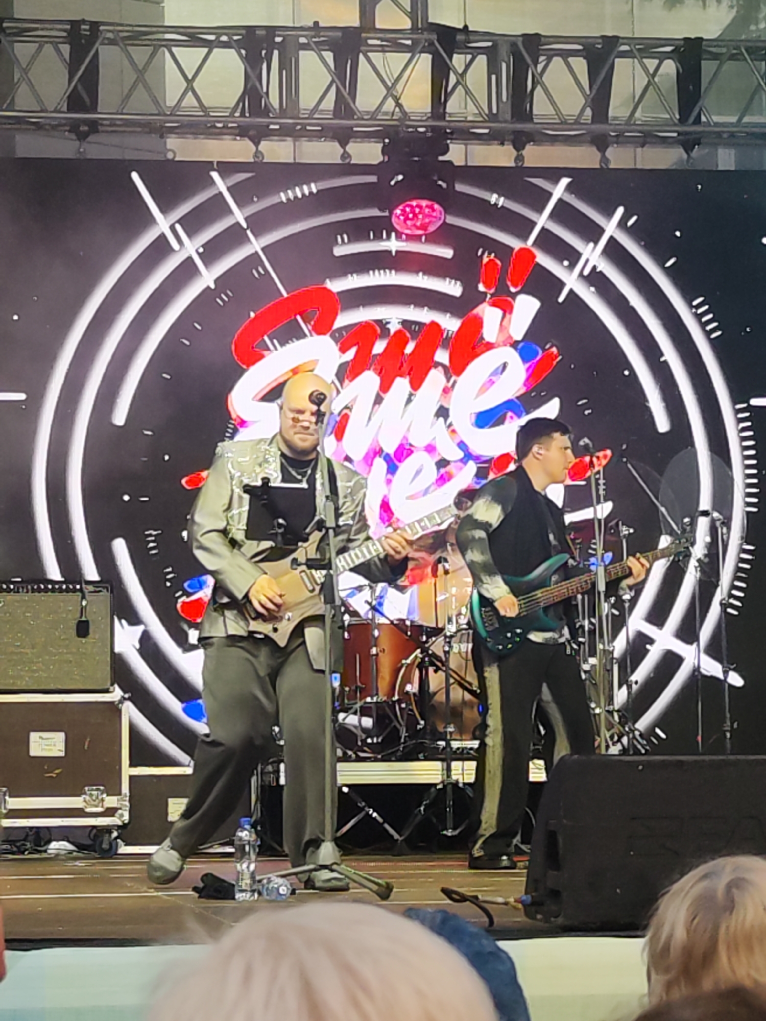 The image captures a live musical performance on a stage, likely in Chaikovsky, Russia, as indicated by the metadata. Two male musicians are the central focus. The musician on the left, who is bald and wearing glasses, plays a light brown electric guitar. He is dressed in a shiny, light grey jacket over a black t-shirt and dark grey pants. To his right, another musician, with dark hair, plays a teal bass guitar. He wears a black jacket with a patterned long-sleeved shirt underneath and dark pants with a stripe down the side. Behind them, a drum set is visible, and a large screen displays a graphic with concentric circles and the word "Sweet" in red and white lettering. The stage floor is visible, with amplifiers, cables, and water bottles on it. The background suggests an outdoor event, with a metal truss structure above the stage. The image is taken from a low angle, looking up at the performers, and the audience is partially visible in the foreground.