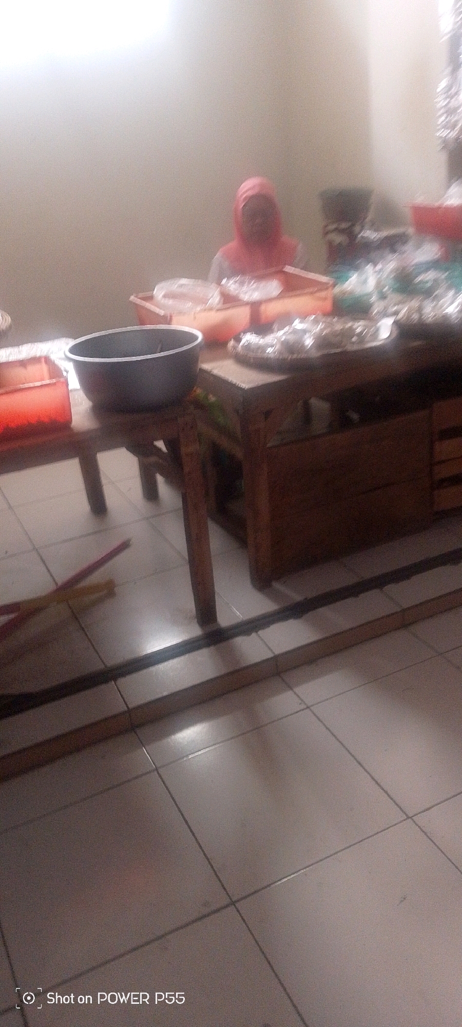 In an indoor setting with tiled flooring, a table and a cabinet are visible. On the table, there are various food items, some wrapped in plastic, and a large dark bowl. A person wearing a pink headscarf is partially visible behind the table, appearing to be engaged in an activity related to the food. The environment is softly lit, with a bright light source creating a hazy effect. There is also a red plastic container on the table and some colorful straws on the floor.