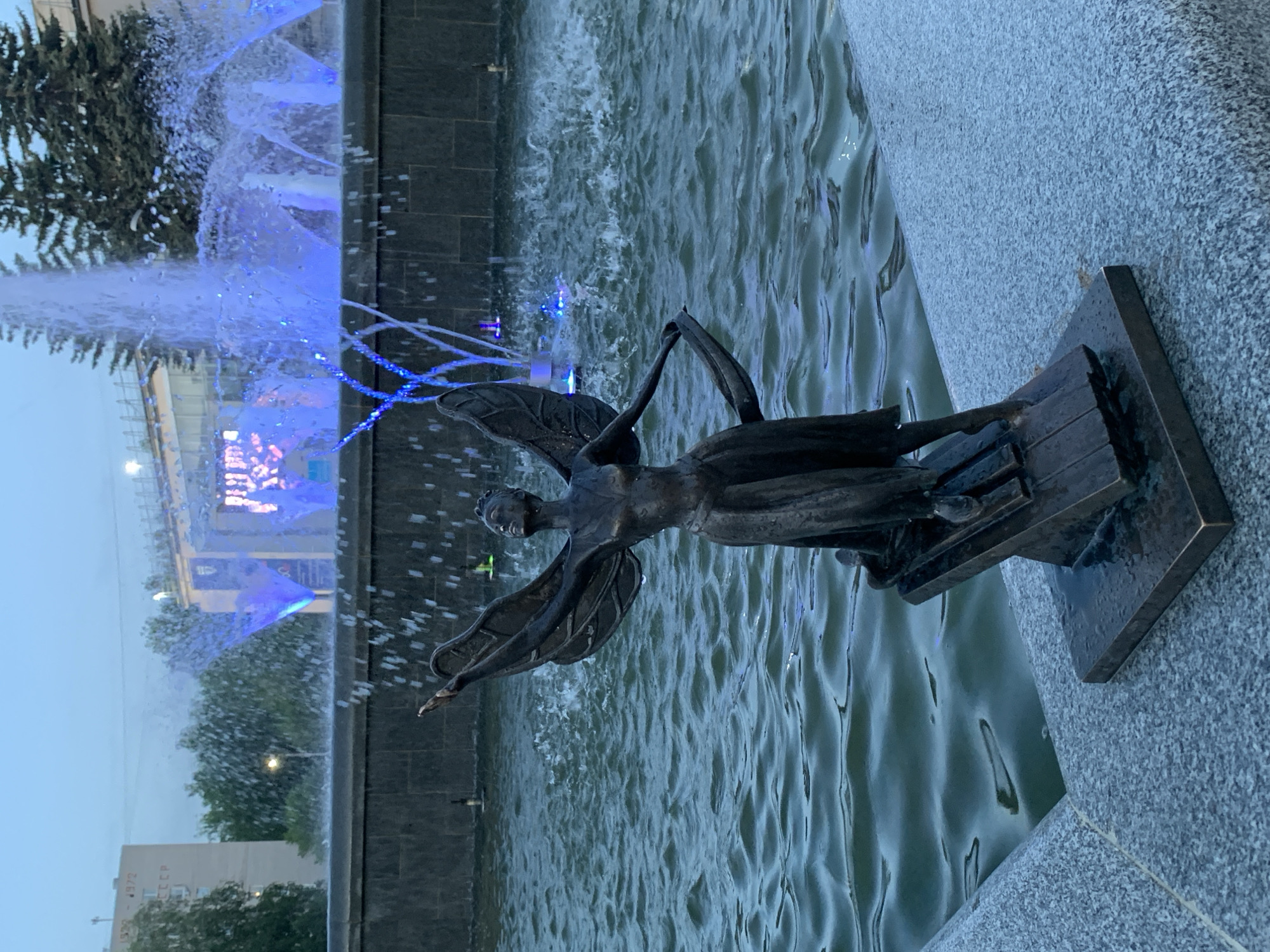 The image captures a bronze statue of an angel, placed within a fountain, likely in the city of Chaikovsky, Russia. The angel, rendered in a darkened bronze tone, is depicted in a dynamic pose with outstretched arms and wings spread as if in flight. It stands on a rectangular, multi-tiered base, also made of bronze. The fountain water surrounds the base and the statue itself.
In the background, water jets are illuminated with a vibrant blue light, creating a visual spectacle and adding depth to the scene. Architectural details of nearby buildings are partially visible behind the fountain, suggesting an urban setting. The statue's placement within the water feature and the lighting effects create a serene yet striking composition.
