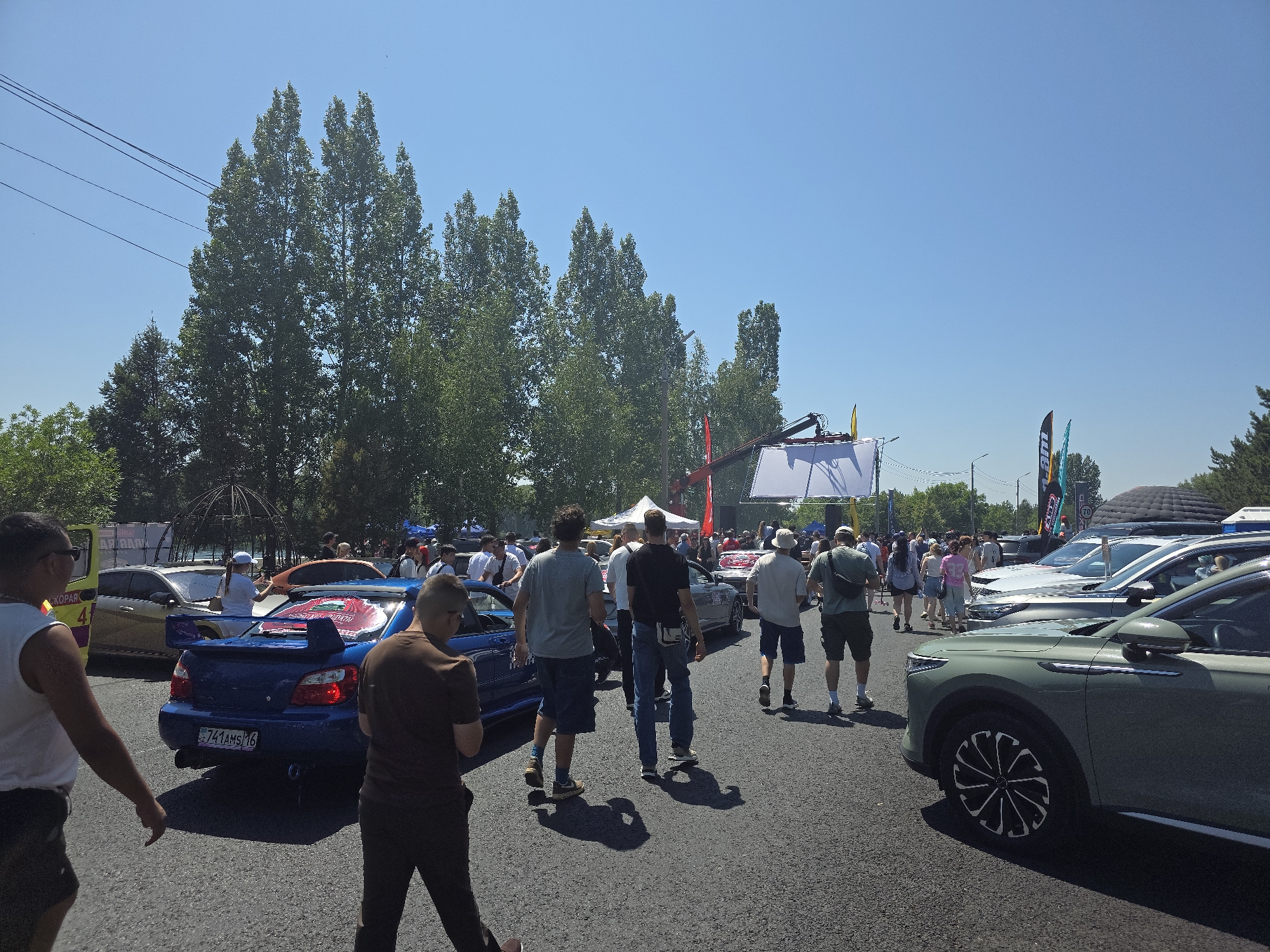 The image captures a sunny outdoor event, likely a car show, in Oskemen, Kazakhstan. A variety of vehicles, including a blue Subaru WRX STI with a prominent spoiler and a green SUV, are parked on an asphalt surface. Several people are walking around, some are looking at the cars, and others are engaged in conversation. In the background, there are trees and banners, suggesting a festive atmosphere. The sky is clear and blue, indicating good weather conditions for the event. The presence of power lines and trees suggests the event is taking place in an open area, possibly a park or a large parking lot within the city.