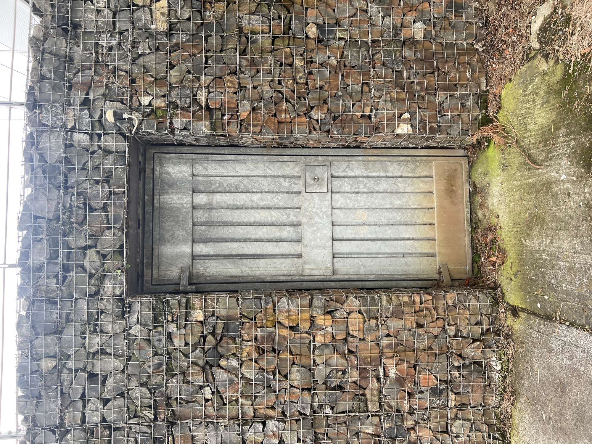 The image depicts a weathered metal door set into a gabion wall. The gabion wall, constructed from wire mesh cages filled with stones, forms the primary structure. The stones are varied in size and color, ranging from dark grey to reddish-brown. The door is grey, with vertical paneling and a horizontal reinforcement bar. A small, circular lock mechanism is visible on the right side of the door. The ground in front of the door is concrete, partially covered with moss and debris such as dry leaves and twigs. The overall scene appears to be outdoors, with a slightly overcast sky visible at the top of the frame. The style of the gabion wall suggests it may be part of a retaining wall or a decorative outdoor feature. No people are present in the image. The context of Balbriggan, Ireland, would imply this is an outdoor structure within a residential, commercial, or public space in that area, possibly a garden boundary, an entrance to a utility area, or part of a landscape design.