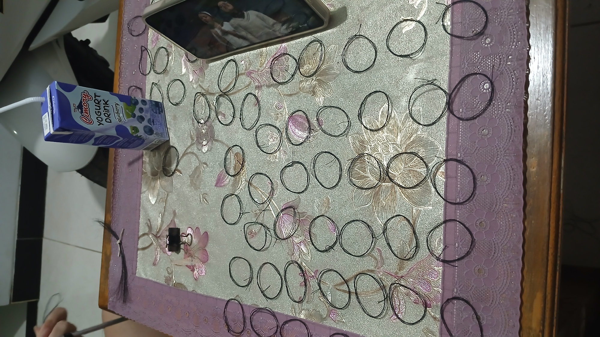 A medium shot captures a flat lay arrangement of numerous small, dark, circular objects, possibly hair ties or wire loops, spread across a patterned tablecloth. The objects are scattered over a floral design on the tablecloth. A purple lace-edged runner lies beneath the tablecloth, at the bottom of the frame. A juice box with the brand "Cimory" and "Yogurt Drink Blueberry" visible is positioned in the upper right quadrant of the frame. A smartphone is partially visible in the lower right, angled towards the viewer, displaying reflections of people. In the upper left, a person's hand is seen holding a silver object, possibly scissors, and manipulating dark strands, suggesting an activity related to the circular objects. The scene is indoors, likely on a wooden table, with a white motorcycle partially visible in the background. This scene is set in Sampang, Indonesia.