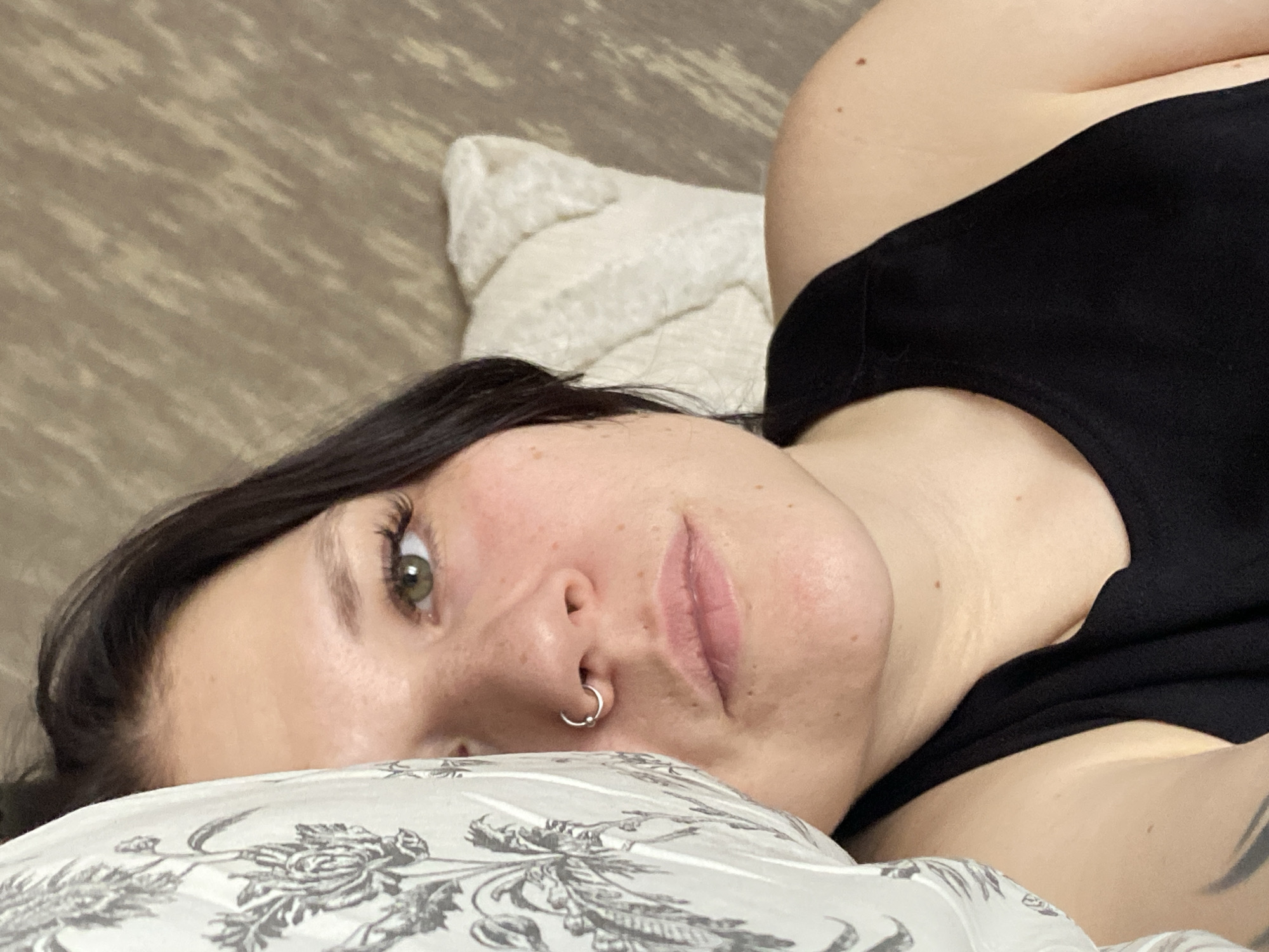 A person with fair skin, dark hair, and green eyes is lying on a bed. The person has a nose ring. The person is wearing a black top. The bed has two pillows, one white and fluffy, the other patterned with gray floral designs. The bed is on a brown surface that might be a headboard. The location is unknown, and there is no indication of Chaikovsky, Russia.