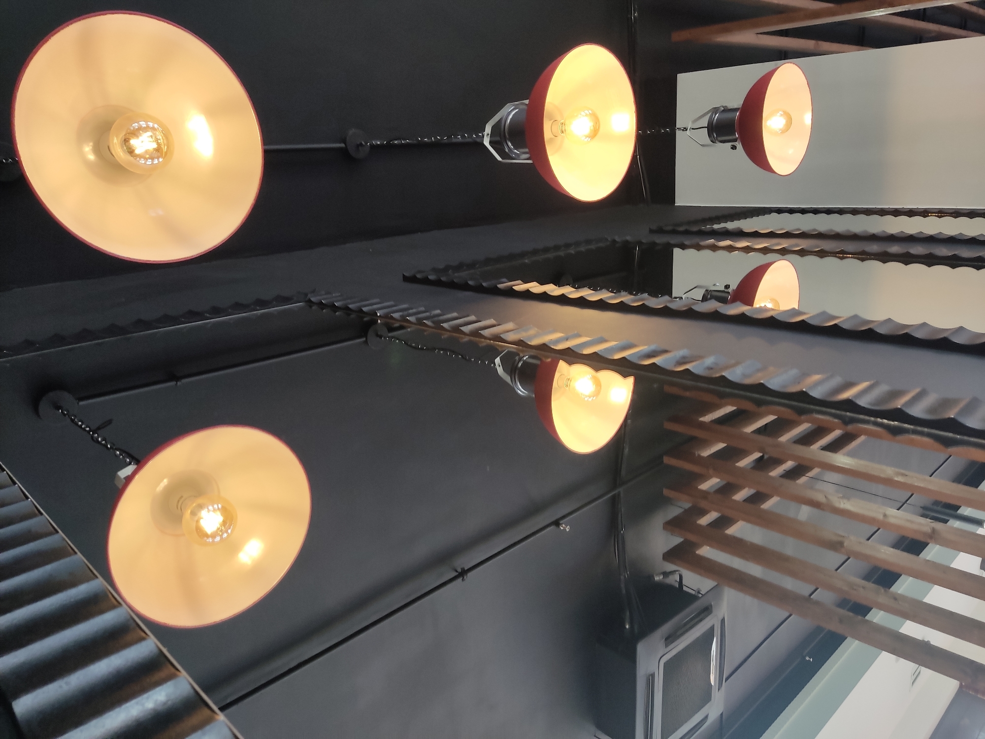 The image depicts a close-up, upward-angle view of multiple industrial-style pendant lights hanging from a dark, textured ceiling. The lights feature red metal shades with a cream-colored interior, illuminating warm-toned incandescent bulbs. Some of the lights are connected by visible chains and electrical wiring. In the background, elements of an interior space are visible, including what appears to be an air conditioning unit, corrugated metal, and wooden slatted partitions or decorative elements. The overall aesthetic suggests a modern, possibly commercial setting like a cafe or restaurant. No people are present in the image, and no specific location in Noginsk, Russia, can be identified.