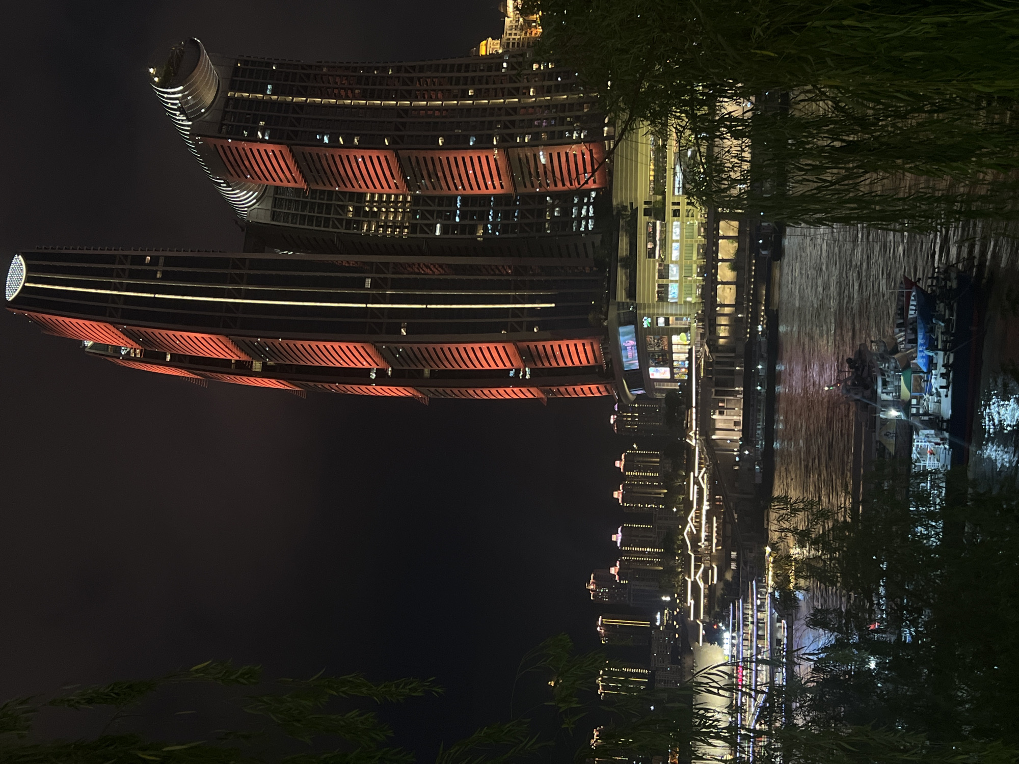 This is a nighttime, eye-level, vertical shot of the Yangtze River waterfront in Jiangbei Cheng Street, China. Two prominent skyscrapers, lit with vertical red and yellow accents, dominate the background. To the left of the skyscrapers, a line of lower buildings with illuminated windows and rooflines stretches along the riverbank. In the foreground, the dark water of the river reflects the city lights. Several boats are visible on the water: a large, multi-deck passenger boat with illuminated windows and railings is docked or moving slowly in the middle distance, and smaller boats are closer to the foreground. The right side of the frame is partially obscured by the dark, leafy branches of a tree, likely a willow, with some of its leaves casting shadows. The sky is dark, indicating night.
