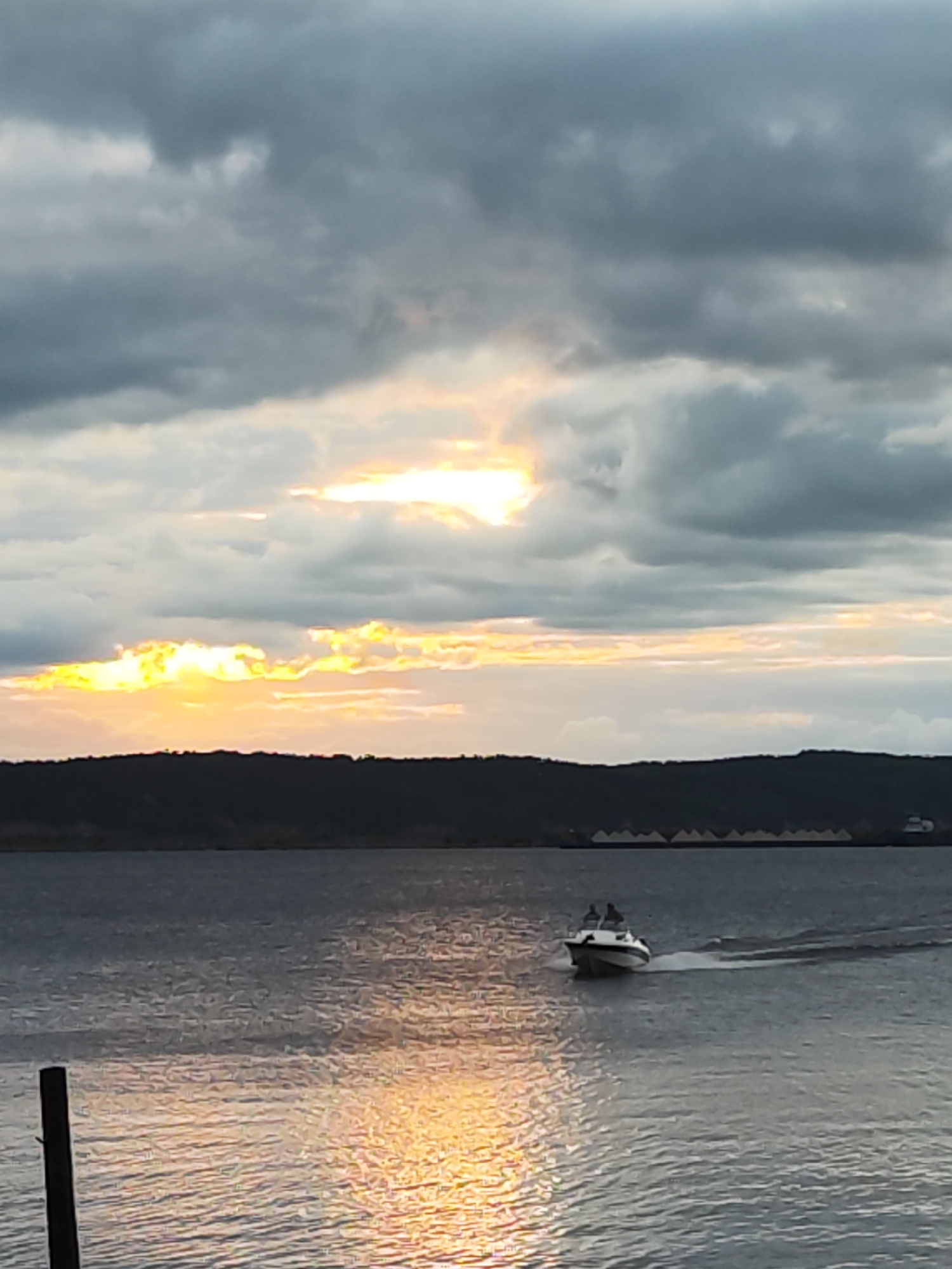 This media file depicts a boat with two people on board moving across a body of water. The scene is set against a backdrop of a dark, tree-lined horizon with a dramatic sky filled with heavy, gray clouds. Bright yellow and orange light breaks through the clouds in the distance, suggesting either a sunrise or sunset. The water is choppy, reflecting the colors of the sky and creating streaks of light on the surface.
In the foreground, a dark wooden post stands vertically on the left side of the frame, partially submerged in the water. The boat, a small white vessel with a dark hull, is in the middle ground, leaving a white wake behind it as it moves from left to right. The two individuals on the boat are small figures, their forms indistinct.
The overall mood is atmospheric and somewhat somber due to the heavy clouds, but the vibrant light breaking through offers a sense of hope or beauty. The time of day is likely dawn or dusk. There is no visible text in the image. The location context provided indicates Chaikovsky, Russia, suggesting a possible river or lake setting within that area.