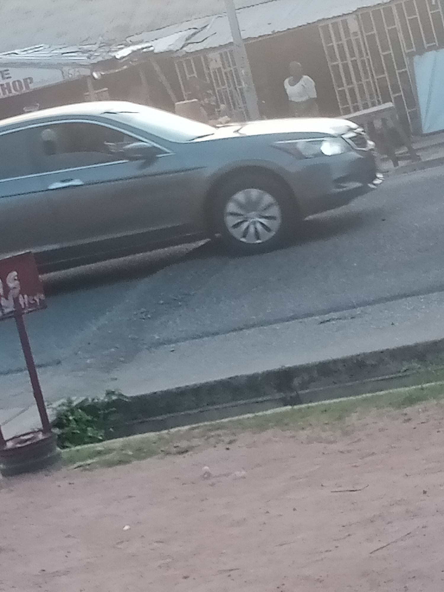 The image depicts a street scene in Jalingo, Nigeria. A dark-colored sedan is parked or driving slowly on the paved road, which appears to be in moderate condition with some cracks and debris. To the left of the car, a red sign with white lettering is partially visible, appearing to be an advertisement or directional marker. Behind the car, a row of structures suggests commercial or residential buildings with corrugated metal roofs and some barred windows. A person in a white top and dark lower garment is visible standing within the doorway of one of these buildings. The foreground of the image shows a dirt or sandy area bordering the road, with some sparse vegetation. The overall lighting suggests it is daytime, possibly late afternoon, given the soft light. The image is slightly blurry, indicating it may have been taken from a moving vehicle or with a less stable camera.