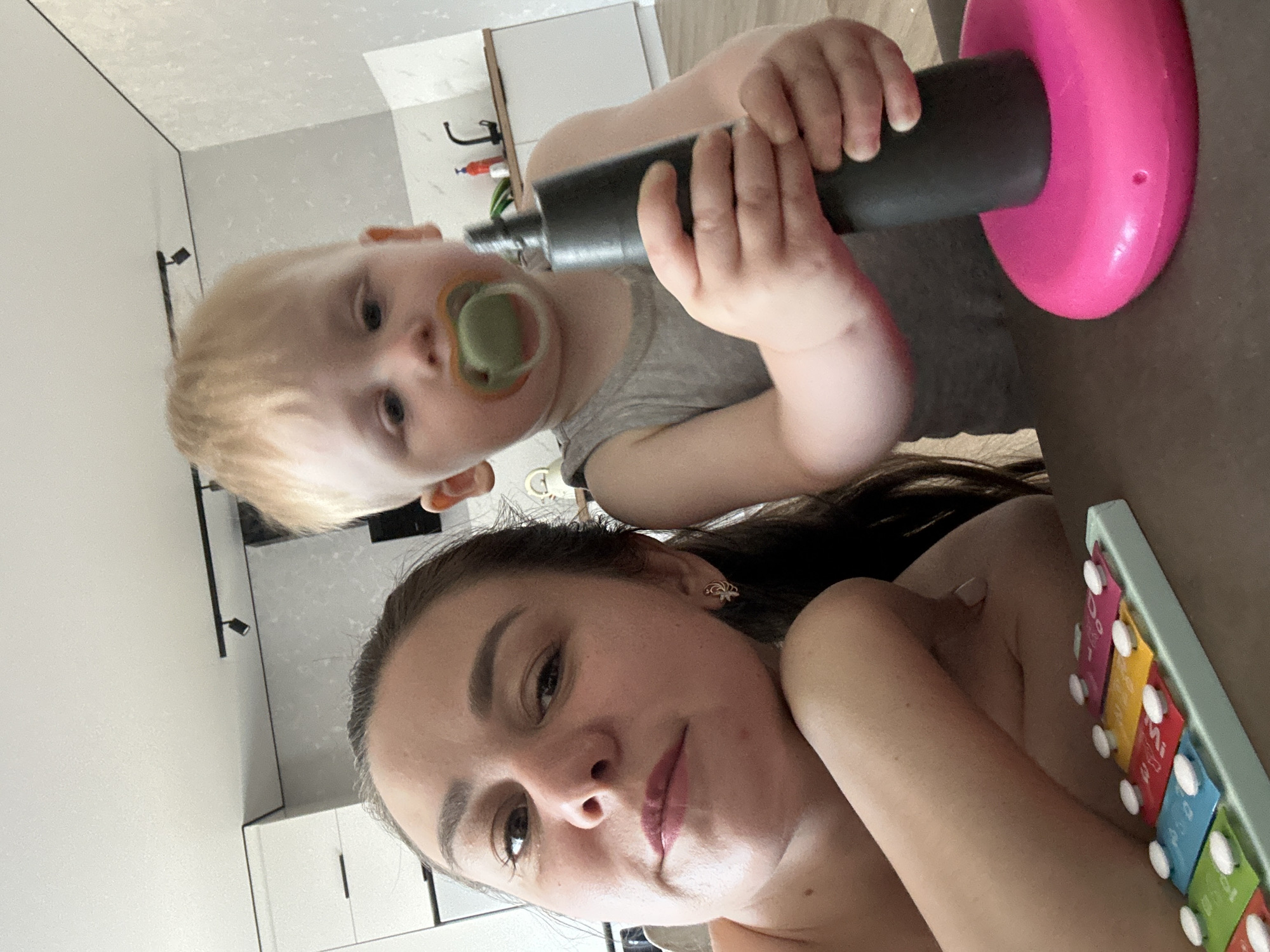 The image is a close-up, selfie-style shot of a woman and a young child in an indoor setting. The woman, on the left, is looking towards the camera with a slight smile. She has dark hair and is wearing a light-colored top. The child, on the right, has blonde hair and a pacifier in their mouth. The child is wearing a grey sleeveless shirt and is holding a black cylindrical object with a pink base. In the foreground, a colorful toy xylophone with blue, green, yellow, and pink keys is visible. The background shows a kitchen area with white cabinets and a faucet. The overall impression is a candid moment of interaction between a mother and child in a domestic environment. The specific location in Chaikovsky, Russia, cannot be determined from the visual information provided in the image.