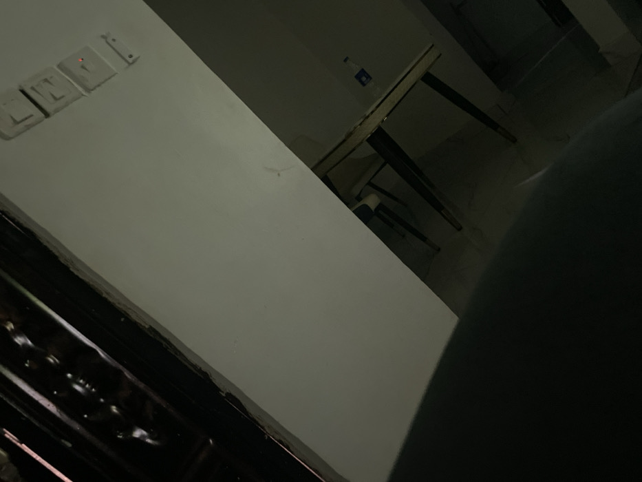 A dimly lit room features a white wall with light switches on the upper left. Next to the wall is a table, also white, with dark-colored legs. A clear bottle is sitting on the table. In the foreground, a large, out-of-focus object obstructs the view. The composition is at an angle, creating a slightly disorienting perspective.