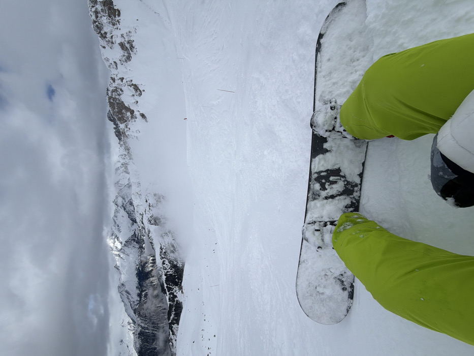 A first-person perspective image depicts a snowboarder resting on a snow-covered piste in Courchevel, France. The foreground shows the snowboarder's legs in bright lime green snow pants, with their feet secured into a black snowboard, partially covered in snow, resting on the ground. Beyond the snowboard, a wide, groomed ski slope descends into a vast mountain range. Distant, rugged snow-capped peaks with exposed rock and scattered evergreen trees are visible under an overcast sky dominated by heavy grey clouds, with some areas partially obscured by atmospheric mist. A red marker flag and thin black piste markers punctuate the slope further down. The scene is a winter mountain environment characterized by extensive snow cover.