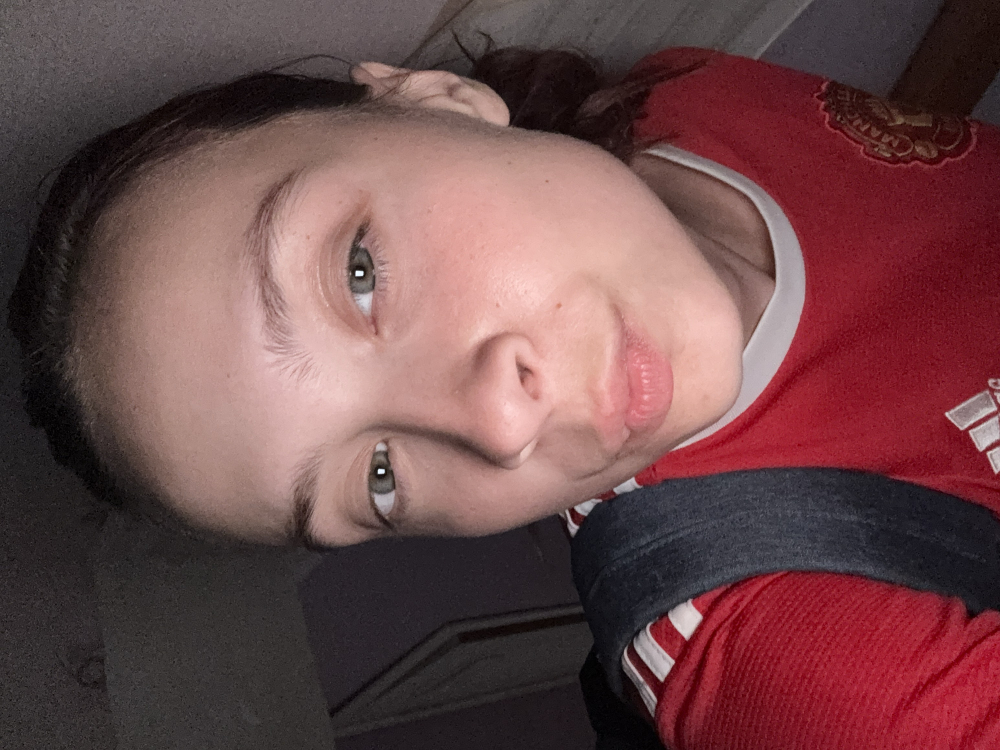 A close-up selfie taken indoors shows a young person with fair skin, light brown hair pulled back, and green eyes. They are wearing a red Manchester United jersey with white stripes on the shoulders and a black backpack strap visible. The person's lips are pursed, and their gaze is directed towards the camera. The lighting is dim, suggesting an indoor environment, possibly a home. The background is dark and slightly blurred, with some indistinct shapes that could be furniture or a wall. There is no visible indication of the specific location within Прикамский, Russia.