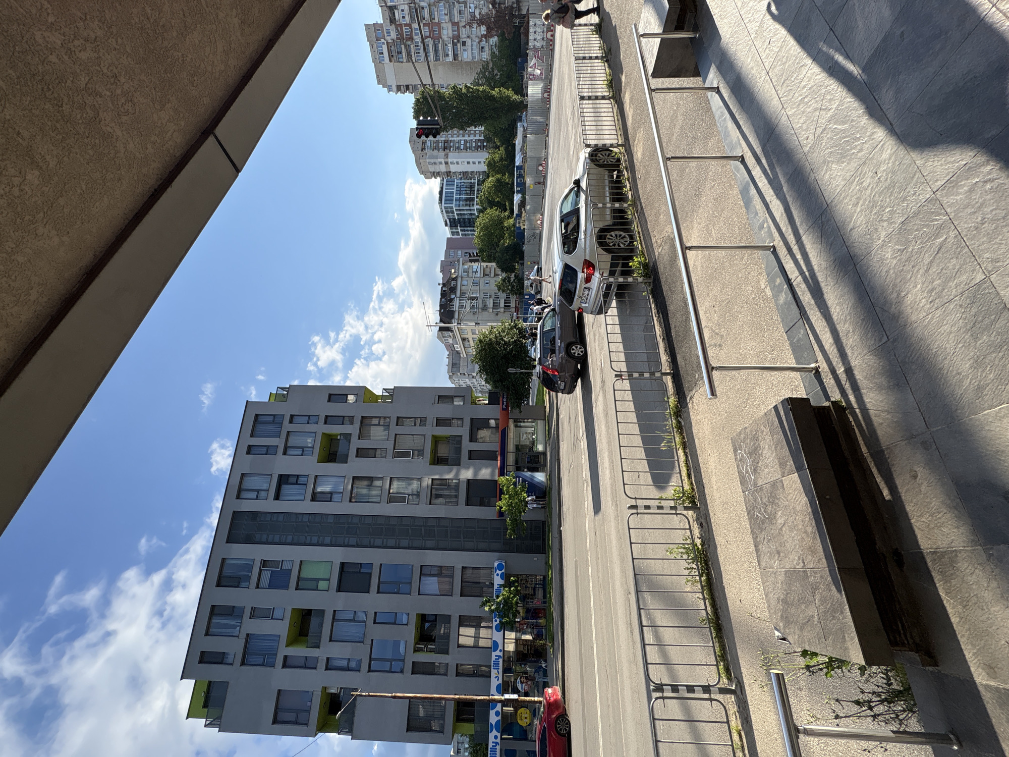 The image shows an urban street scene in Sofia, Bulgaria. The perspective is looking out from a building. The sky is blue with scattered clouds. A modern grey building with multi-colored accents sits opposite the viewpoint. The street has parked cars, a traffic light, and a metal fence running along the sidewalk. A woman is walking along the sidewalk in the background. There's a row of older apartment buildings further down the street. The shadow of the photographer or the building they are in is visible in the bottom right.