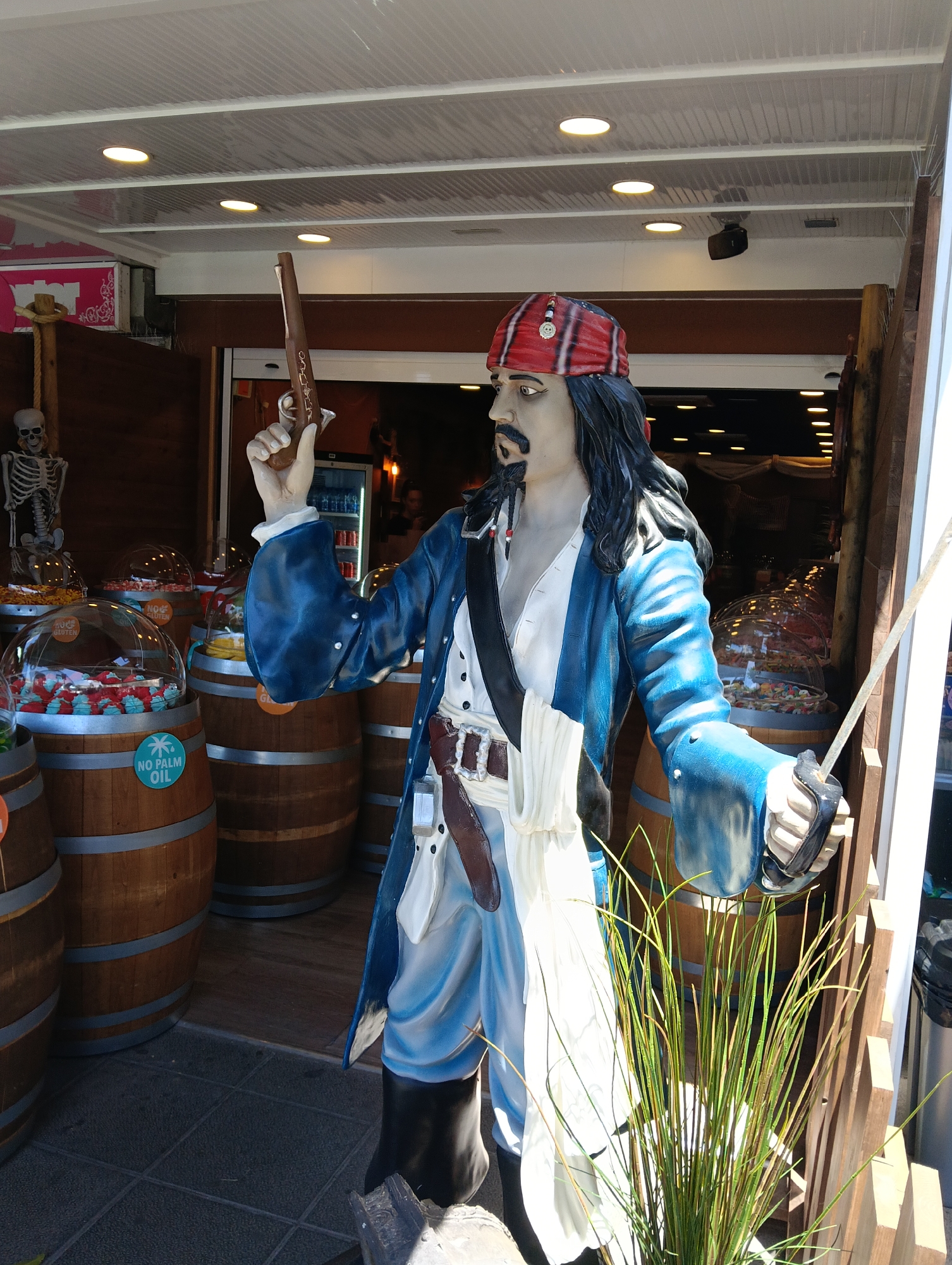 A life-sized statue of a pirate, strongly resembling Captain Jack Sparrow, stands prominently at the entrance of a candy or souvenir shop. The statue is the central subject, dressed in a red and black striped bandana, a white shirt, a blue coat, a brown belt, and black boots. It holds a long, brown flintlock pistol in its raised right hand and the hilt of a sword with a rope-like blade in its left.
The setting is an open-fronted shop, featuring a pirate theme. In the background, several wooden barrels with silver bands serve as display units, each topped with clear, domed containers filled with colorful sweets. One barrel clearly displays a round teal sticker with a palm tree icon and the text "NO PALM OIL". Other candy domes show partially visible text like "NO GLUTEN". To the far left, a white plastic skeleton hangs from a wooden post, adding to the themed decor.
The scene is brightly lit, suggesting a sunny daytime. The ceiling above is white and corrugated, featuring several recessed circular lights. The foreground has a dark grey tiled floor, transitioning to a wooden floor inside the shop. A cluster of green, reedy plants is visible in the lower right. The pistol appears to have decorative carving. The overall atmosphere is playful and inviting, typical of a tourist-oriented establishment in a location like Puerto Del Carmen, Spain.