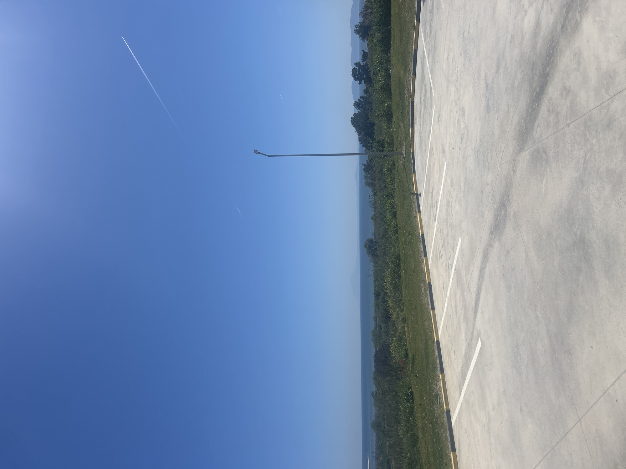 The image depicts a parking lot next to a grassy area with trees, likely located near Νέα Κερδύλια, Greece. The sky is blue with airplane contrails. A tall light post is present near the edge of the parking lot and the grassy area. The parking lot surface is concrete with painted white lines and yellow and black barriers. In the background, there are hills or mountains. The weather is sunny. No people are visible.