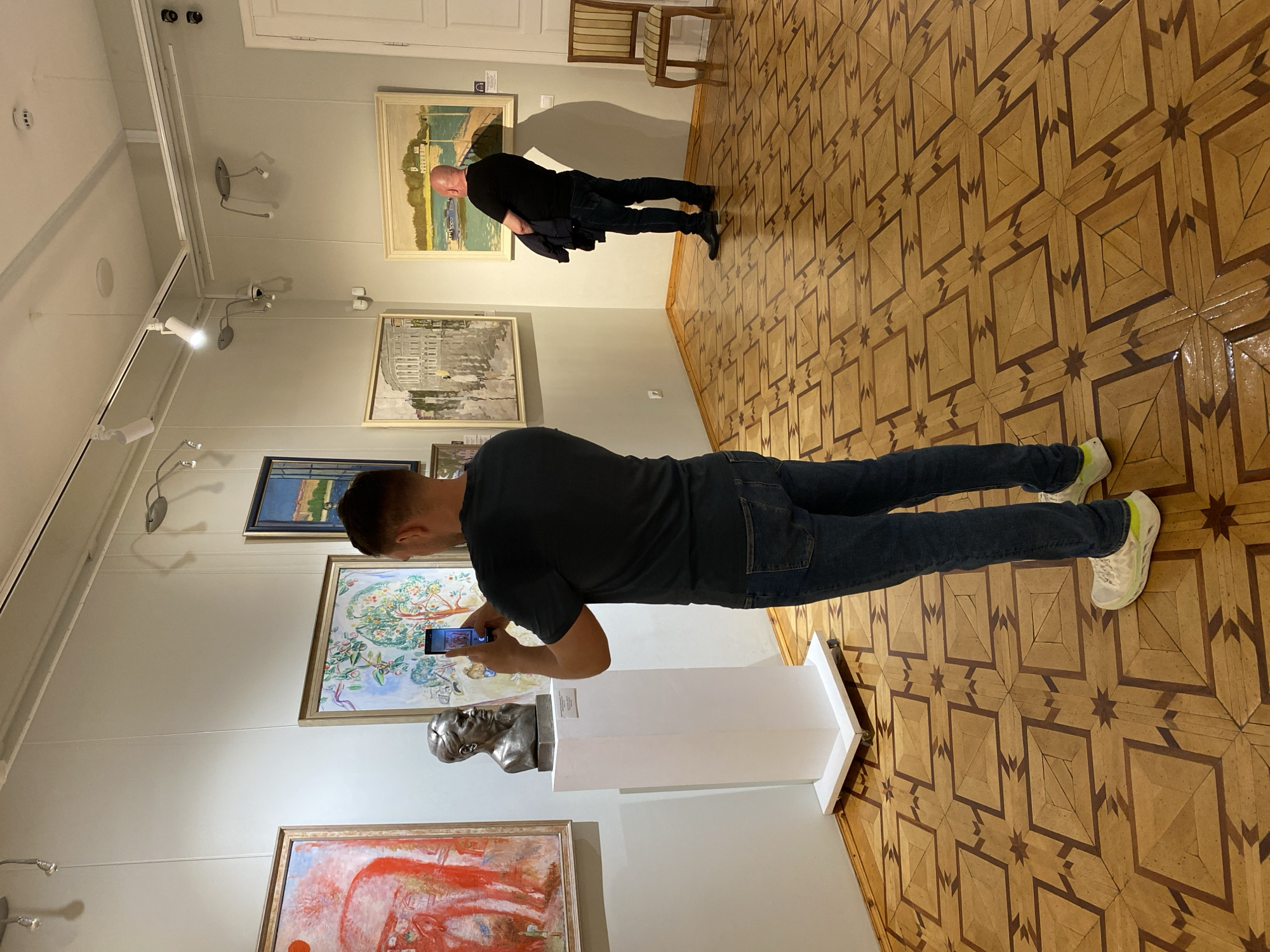 The image shows the interior of an art gallery, possibly the National Gallery of the Republic of Karelia in Petrozavodsk, Russia, judging by the context of other images from this collection. Two men are present. In the foreground, a man with dark hair is standing and holding a smartphone, taking a picture of a bronze bust displayed on a white pedestal. The bust appears to be of a man with short, slicked-back hair and a stern expression. Behind this man, another man with a shaved head is standing, facing away from the camera, wearing a black t-shirt and dark jeans. He is holding a dark jacket over his left arm and looking at paintings on the wall. The gallery walls are a neutral color and display several framed artworks, including a large abstract painting with red and white elements on the left, and other landscape or cityscape paintings further back. The floor is made of polished hardwood with a distinctive geometric inlaid pattern. Lighting in the gallery is provided by track lighting mounted on the ceiling. A single wooden chair with a striped seat and back is visible on the right side of the frame.