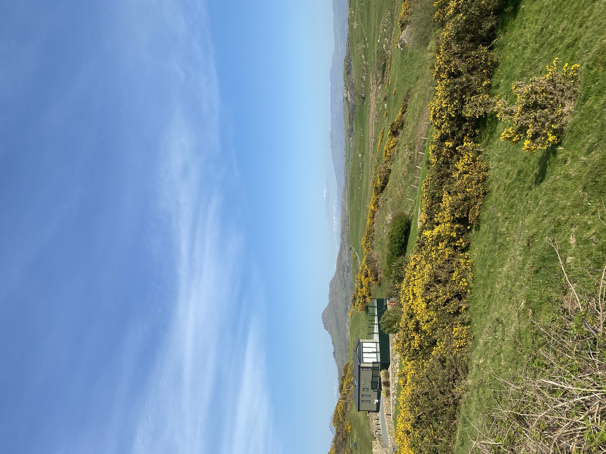 The image shows a coastal scene in Criccieth, United Kingdom. The sky is a bright blue with whispy clouds. The landscape features green hillsides dotted with bright yellow gorse bushes. A modern cabin-style building with a dark green exterior sits on the slope, partially obscured by the gorse. In the background, hazy mountains are visible.