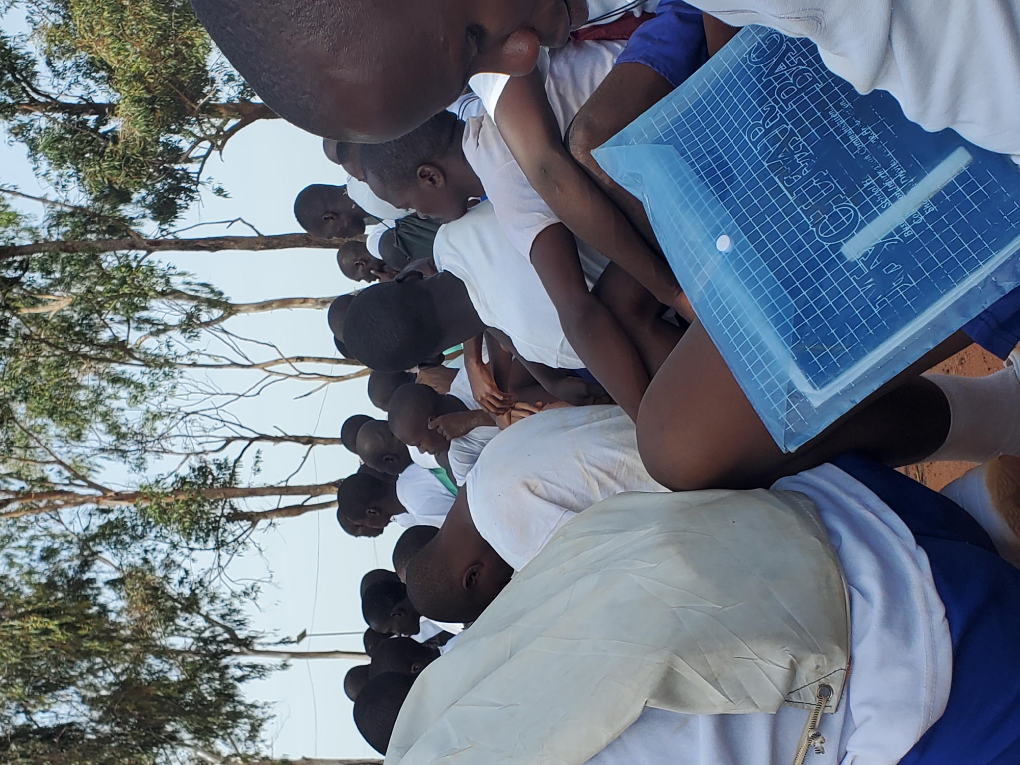 In the city of Barakin Rafin Gora, Nigeria, a group of young people, mostly boys, are gathered outdoors under tall trees with sparse foliage against a clear sky. They are dressed in white tops, with some wearing blue shorts. The individuals are in close proximity, suggesting a communal gathering or event. The foreground is dominated by a close-up of a person in a white t-shirt and blue shorts, holding a blue transparent plastic folder. Another person's head and shoulder, also in a white top, are visible to the left. The folder displays text which includes "CLEAR BAG" and other smaller inscriptions that are partially legible, mentioning "School of Information and Communication" and "Business College," hinting at an educational context. The overall scene appears to be one of a gathering for a school-related activity or event.