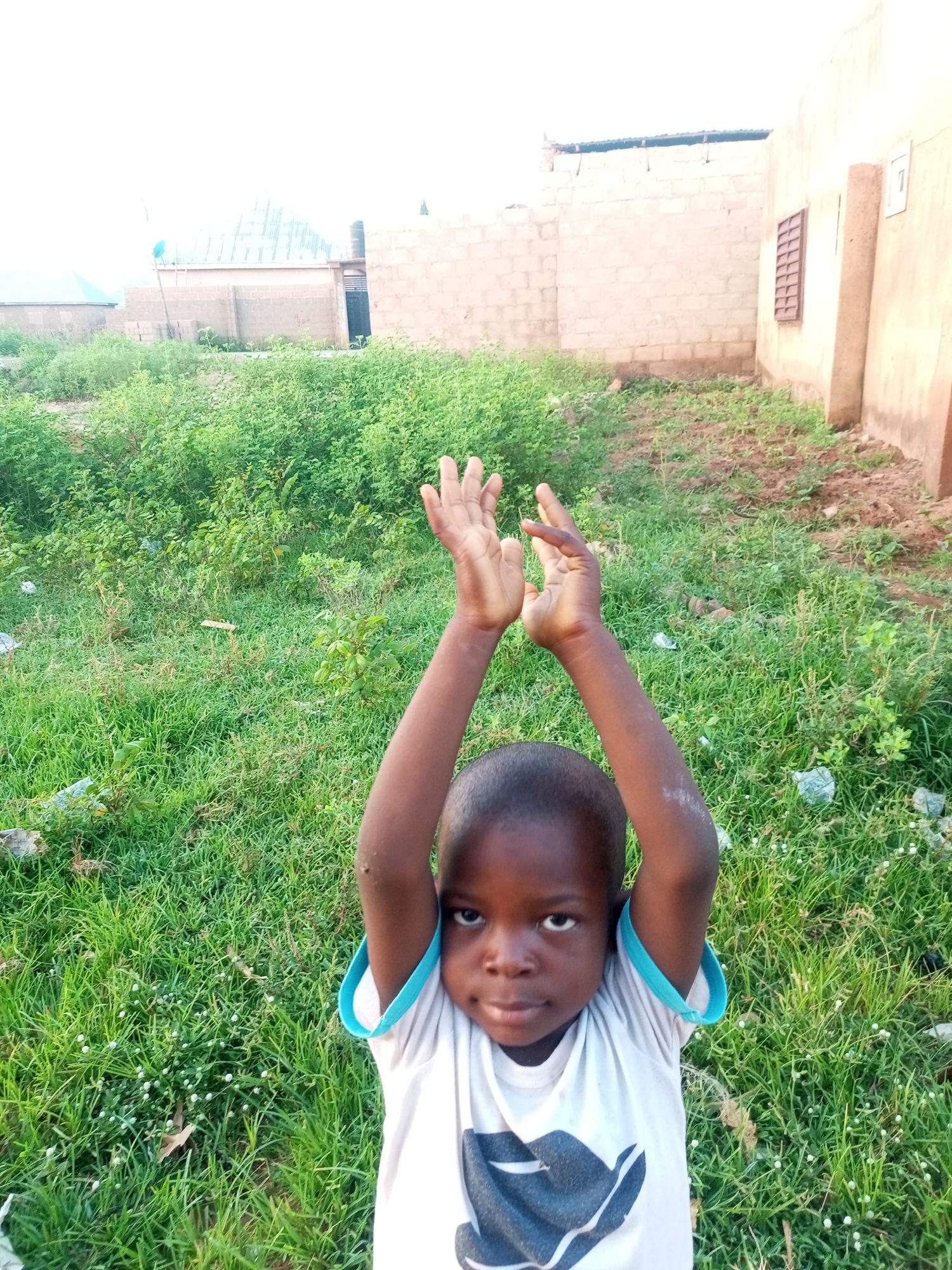 The image depicts a young child with dark skin and hair, wearing a white t-shirt with a blue trim. The child's arms are raised, and their hands are open, held at chest level. The child is looking directly at the camera with a neutral expression. The background consists of a grassy area with some scattered debris and buildings in the distance. The buildings appear to be constructed from bricks and have various roofing materials. The environment suggests an outdoor setting, possibly in a residential area of Tempure, Nigeria. The lighting suggests it is daytime, with soft light conditions.