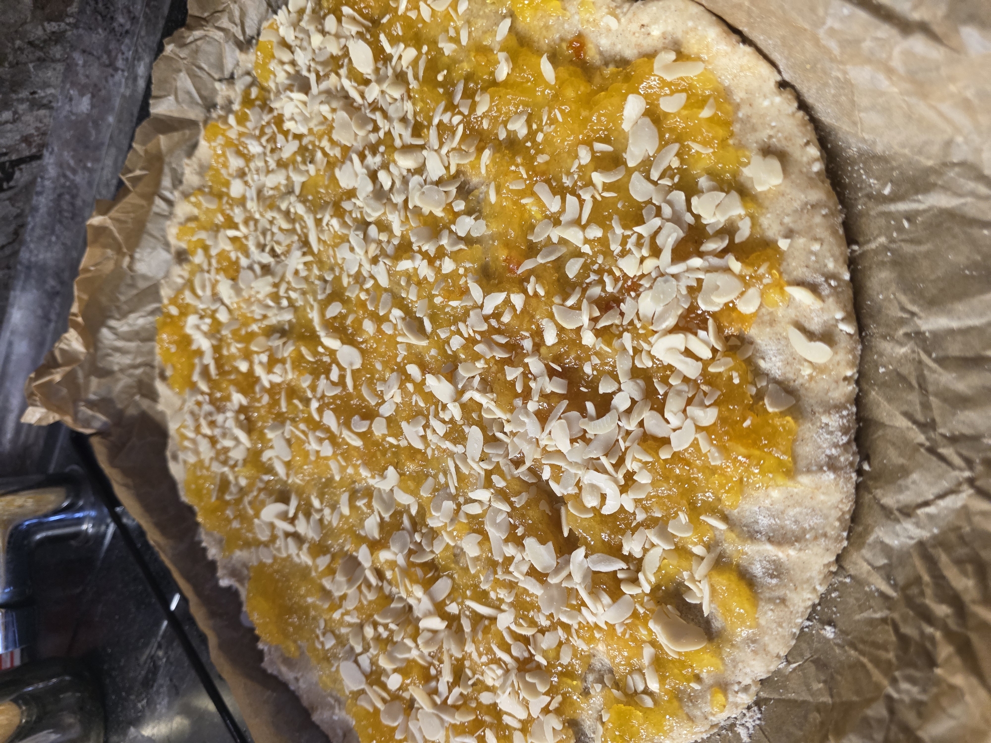 An overhead, close-up shot shows a whole wheat flatbread topped with a bright yellow jam and slivered almonds. The flatbread is on a baking sheet lined with parchment paper, which is then placed on a countertop. In the background, a stainless steel faucet is visible, suggesting the flatbread is in a kitchen. The lighting is natural and coming from the right side of the frame. There are no people or animals in the image.