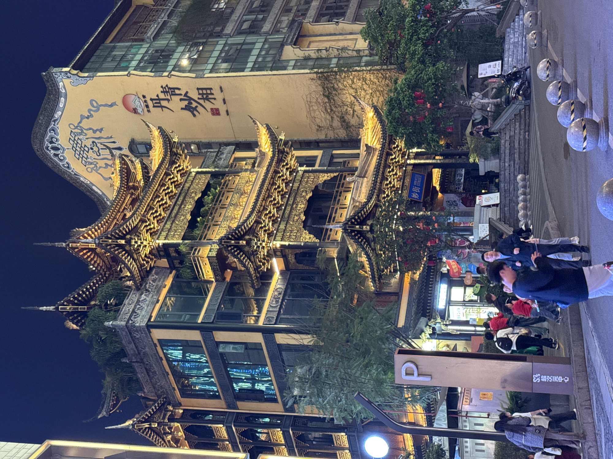 The image captures a night scene in the city of Yuzhong District, China. An ornate building with traditional Chinese architectural elements, including tiered roofs and intricate carvings, dominates the left side of the frame. It's illuminated, highlighting the details of the structure. To the right of the building, pedestrians stroll along the street. A tall parking sign is visible in the lower right corner. The street is lit by street lamps. In the background, there are other buildings and structures that contribute to the urban landscape.