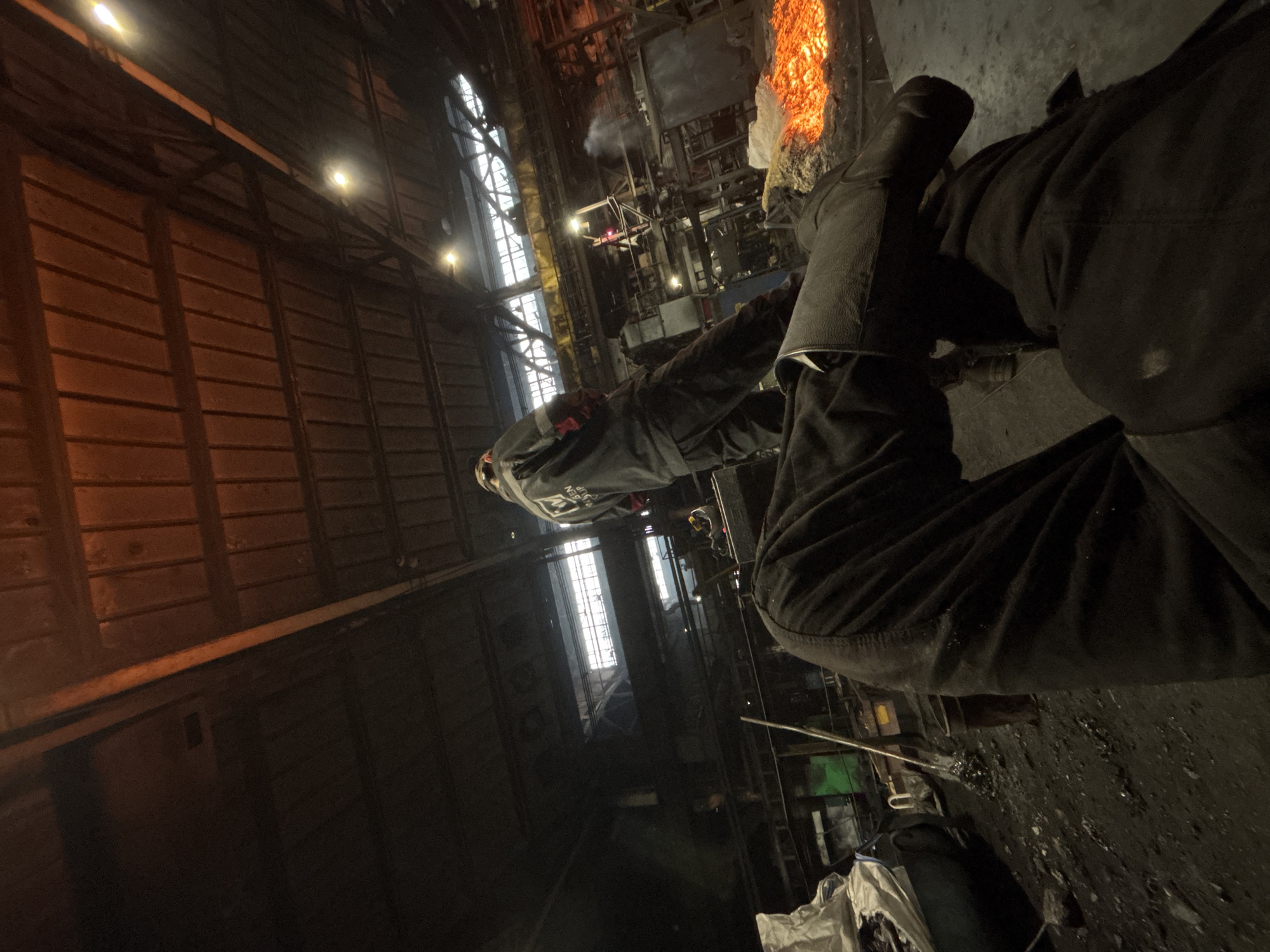 The image depicts an industrial setting, likely a foundry or workshop, characterized by dim lighting and a grimy atmosphere. In the foreground, the lower body of an individual wearing dark work pants and heavy boots is visible, suggesting they are seated or in a relaxed posture. Further into the scene, another person is standing, dressed in a dark work jacket with reflective detailing and a cap, facing away from the camera. The environment is filled with industrial equipment and materials. A prominent element is a large pile of glowing, molten material, emitting a bright orange light and heat. Scattered around are various tools, likely for handling the molten material, including a shovel. The overall impression is one of an active, hot industrial workspace. The specific location in Homyel, Belarus cannot be determined from the visual information alone.