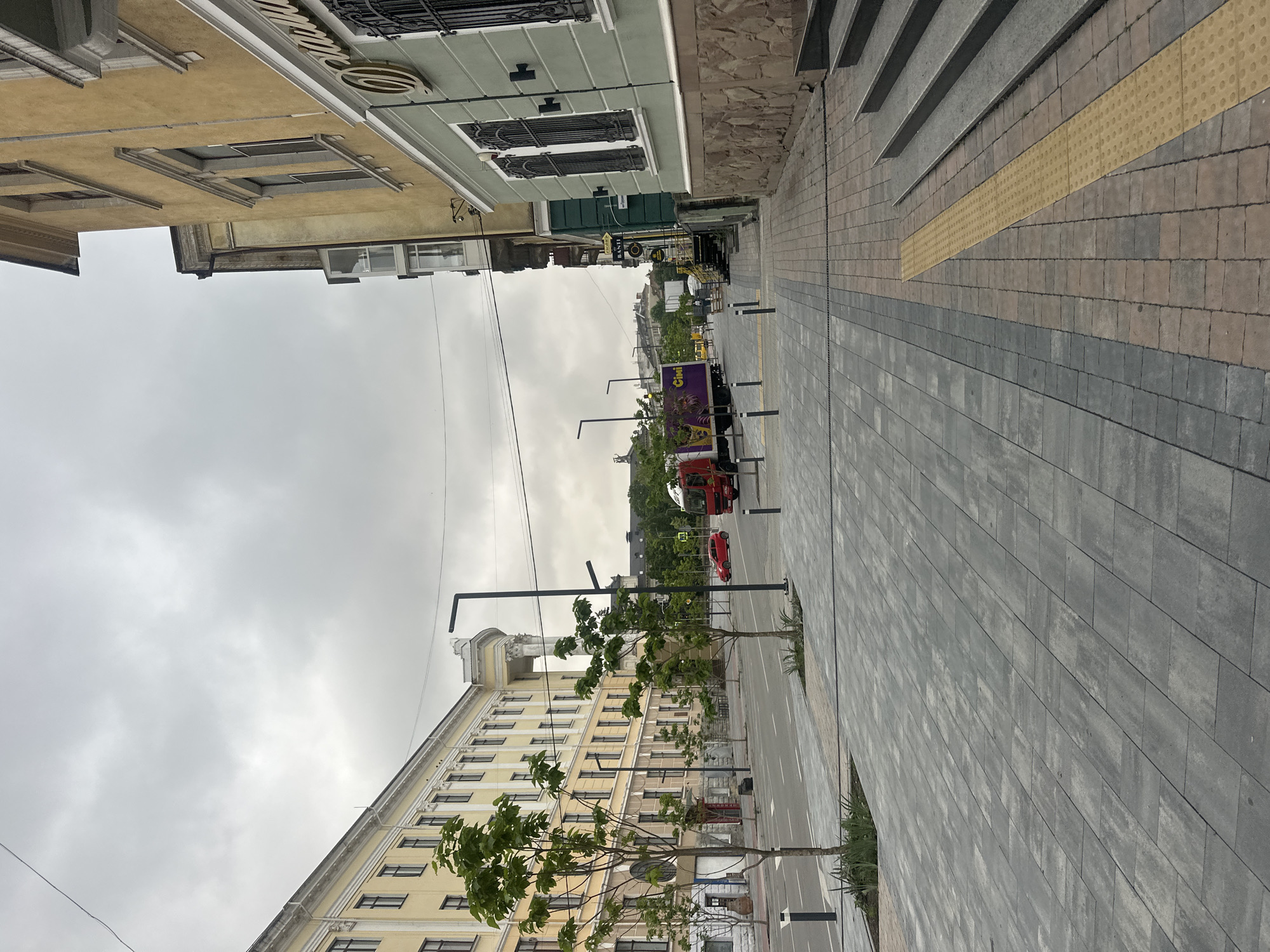 This image depicts a street scene in Ternopil, Ukraine, under an overcast sky. A wide sidewalk paved with grey rectangular tiles lines the right side of the image, featuring a yellow tactile paving strip adjacent to a set of concrete stairs. To the left, a street runs parallel to the sidewalk, with a red car visible in the distance. On the left side of the street stands a large, light-yellow building with numerous windows and classical architectural details. A row of young trees with green foliage is planted along the sidewalk in front of this building. Across the street, a red truck with a purple and yellow graphic on its side is parked, and further down the street, other buildings and trees are visible. The scene is devoid of people and animals.