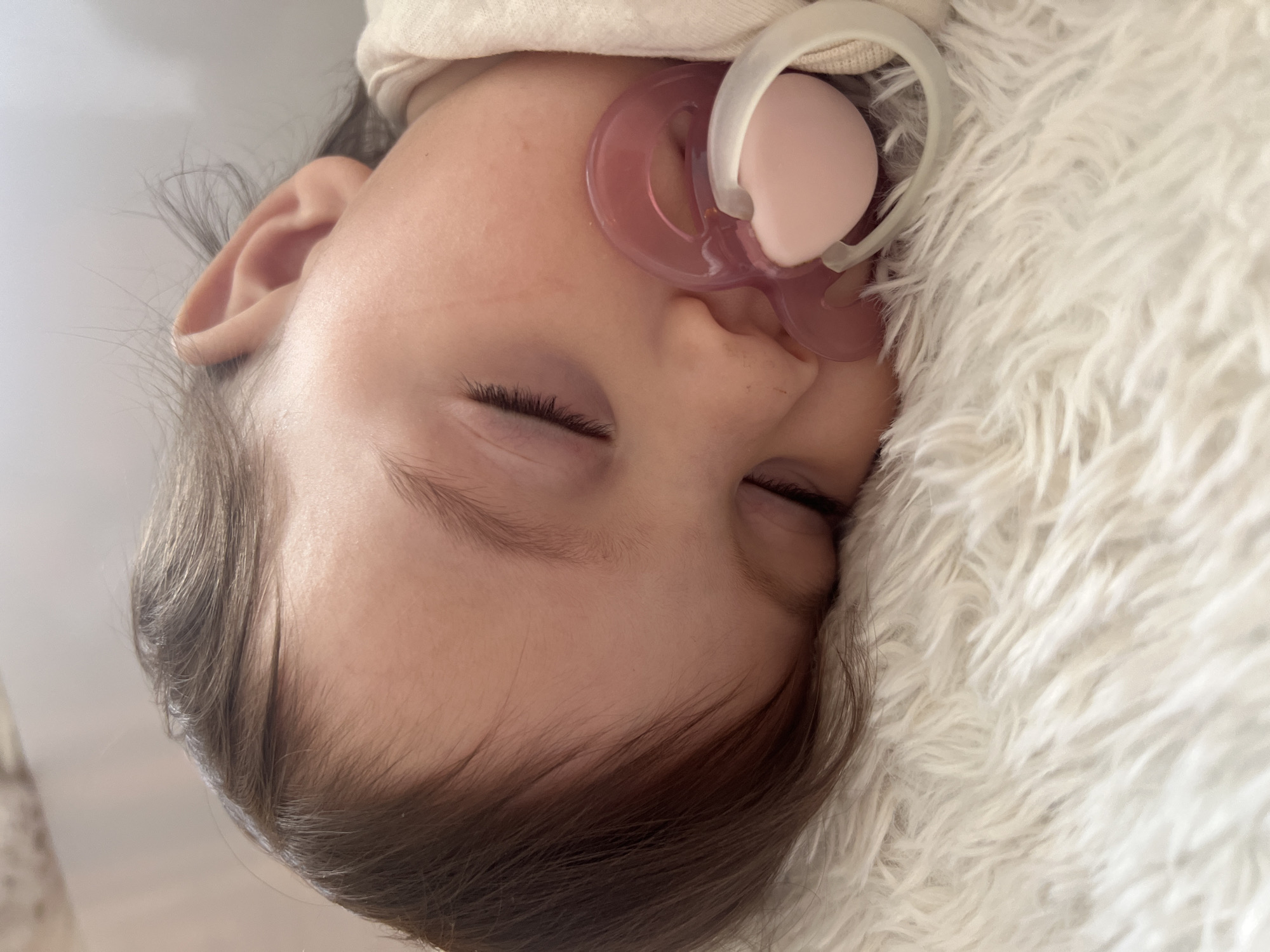 A close-up, upside-down image shows a baby asleep, with a pink pacifier in its mouth. The baby's face is fair, and its eyes are closed with visible eyelashes. Dark brown hair is visible around the forehead. The baby is lying on a white, soft, and fluffy material. No elements indicate the location is in the city of Elena, Bulgaria.