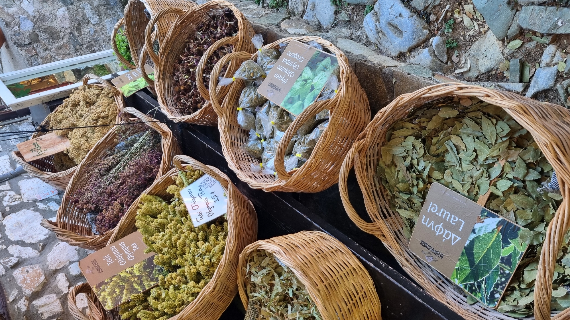 This is a close-up, top-down shot of several woven baskets filled with dried herbs and spices, likely at an outdoor market stall. The baskets are arranged in a tiered fashion, with some spilling over the edges. The herbs vary in color and texture, ranging from fine, light yellow clusters to darker, more stringy purple and brown varieties, as well as a basket full of green leaves.
One basket prominently displays small, individually packaged bags of dried herbs, with a label indicating "Olympus Oregano". Another basket contains a large quantity of dried green leaves, identified by a label as "Laurel". In the foreground, a basket is filled with small, light green, flower-like dried herbs, with a label reading "Olympus Tea" and a price of "1.5€". The ground beneath the baskets is paved with irregular stones.
The scene suggests a rustic, traditional setting, consistent with a village market in Greece, as indicated by the Greek text on the labels. The lighting suggests it is daytime, and the overall impression is one of abundance and natural products. The text visible on the labels includes product names like "Oregano", "Laurel", and "Olympus Tea", along with brand information such as "Olympussenses".