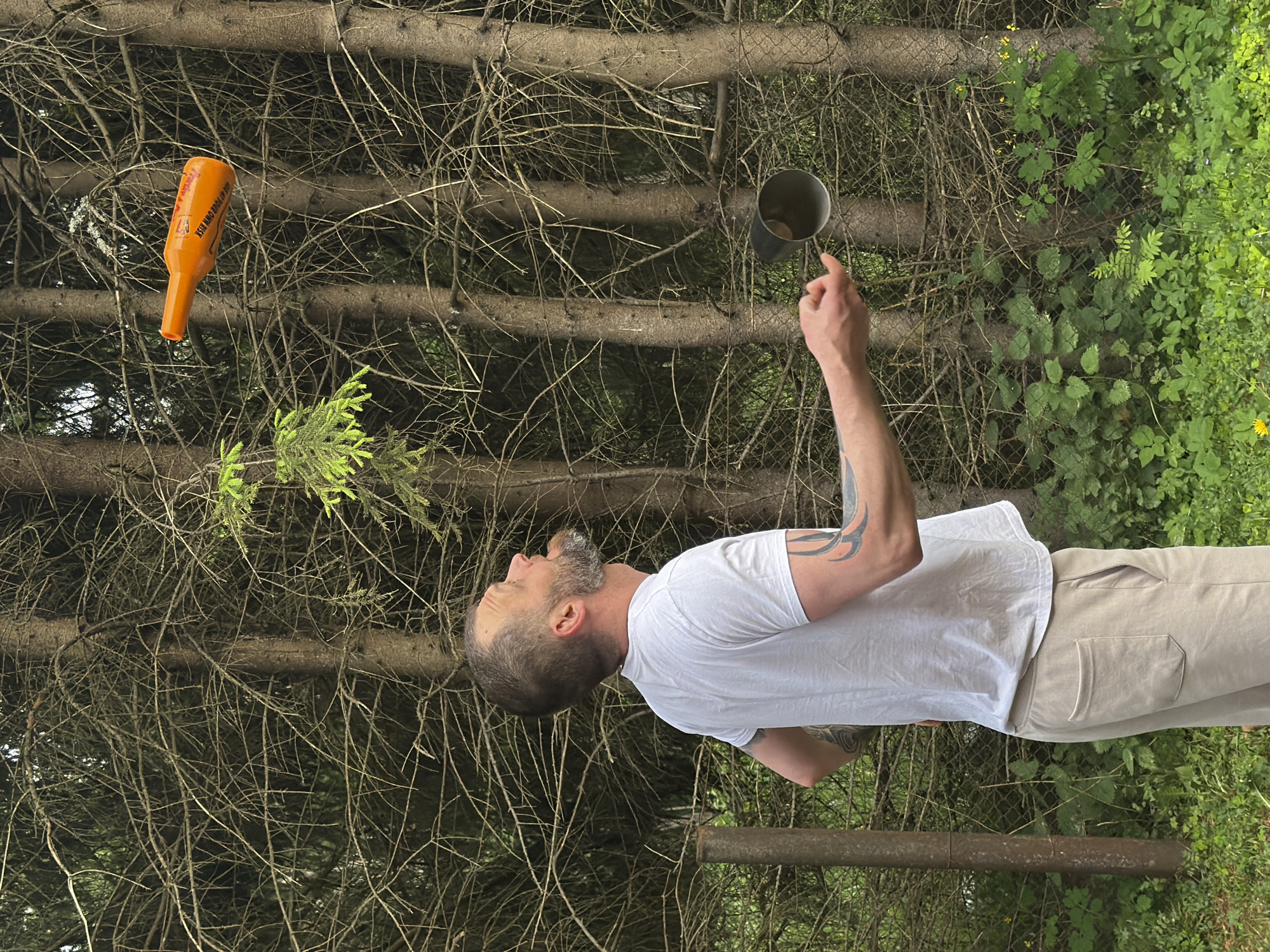 An adult male with a beard and tattoos on his arms juggles an orange bottle and a metal cup outdoors. He is wearing a white t-shirt and beige pants. The background consists of a dense forest with numerous trees and green foliage. The scene appears to be a park or wooded area within Tallinn, Estonia.