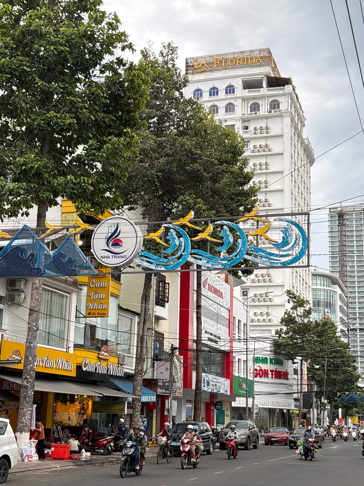 The image shows a street scene in Nha Trang, Vietnam. In the foreground, a street is lined with buildings and businesses. The street is busy with traffic, including motorcycles, cars, and bicycles. People are seen riding motorcycles, walking, and engaging in various activities.
One prominent building in the background is a tall white building with the word "FLORIDA" in orange lettering at the top. The building has multiple floors with arched windows and several air conditioning units mounted on its facade.
In the middle ground, there are several businesses with signs. One sign reads "Com Tam Nhan" which is a popular Vietnamese dish. Another sign is for "HONDA". There's also a circular sign with "NHA TRANG" and a logo depicting waves and a sunset, which is a common symbol for the city.
On the street, there are numerous motorcycles and a few cars navigating the road. Some motorcycles are carrying multiple passengers. Pedestrians are visible on the sidewalks and near the buildings. Trees with dense green foliage are interspersed along the street, providing shade. The sky is overcast, indicating a cloudy day. The overall scene captures the dynamic and lively atmosphere of a street in Nha Trang.