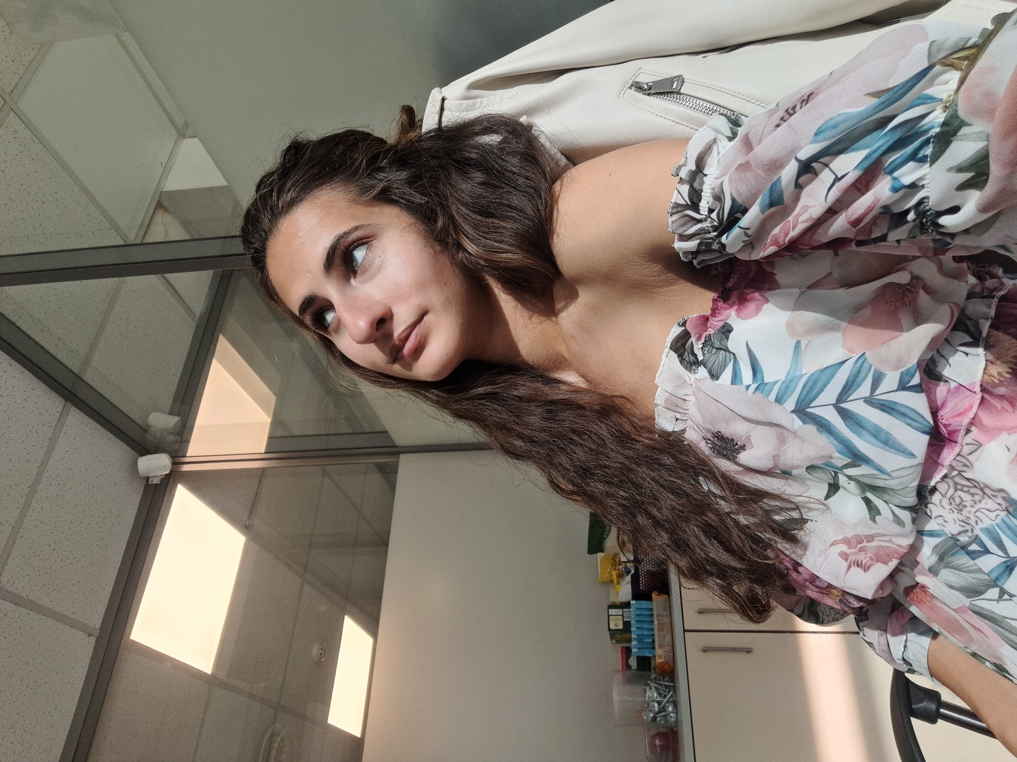 A young woman with long, dark brown hair is looking upwards. She is wearing an off-the-shoulder floral print dress with pink and blue accents. A white leather jacket is draped over her right shoulder. She is seated in a chair in what appears to be an office environment, with glass partitions, suspended ceiling tiles, and fluorescent lighting visible. A shelf with various items, including what appear to be screws or nails in a container, is in the background. The sun is casting a shadow across her face and chest. The scene is lit by natural sunlight and overhead artificial lighting. The image was taken indoors, likely in an office building. The location of Sofia, Bulgaria, is not discernible from the visual information provided in the image.
