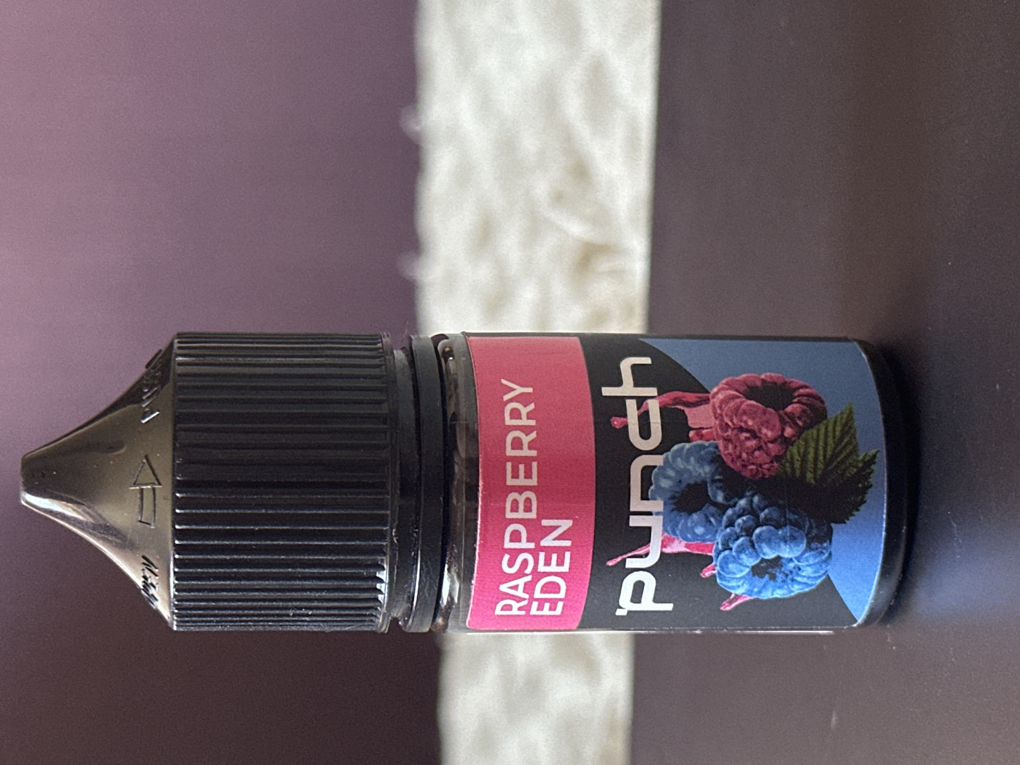 A close-up, eye-level shot captures a bottle of e-liquid with a black cap and a pink and black label featuring the text "RASPBERRY EDEN PUNCH" and an illustration of raspberries. The bottle is placed on a dark, reflective surface, likely a table, with a blurred, fuzzy white object and a purple background in the distance. There are no people or actions depicted in the image. The scene is indoors and does not provide any information about the city of Uzhhorod, Ukraine, or any specific location within it.