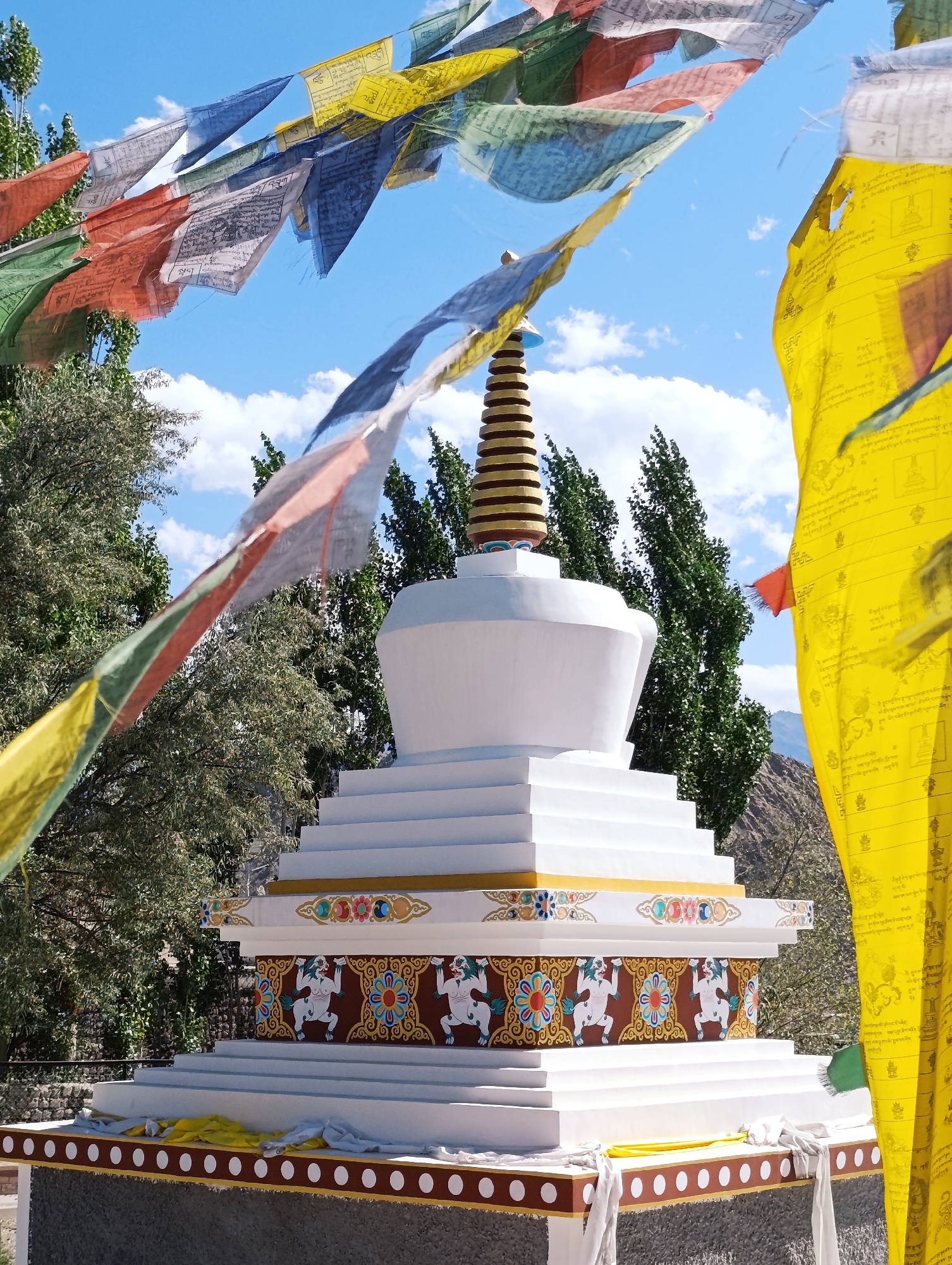 The image depicts a white stupa adorned with colorful decorative bands in the city of Leh, India. Prayer flags in various colors, including blue, yellow, red, green, and white, are strung across the foreground and middle ground, fluttering in the wind against a bright blue sky with scattered clouds. Trees with dense green foliage frame the stupa on the left and in the background. The stupa itself has a tiered base with intricate patterns and motifs, including depictions of mythical creatures and floral designs. A conical finial with golden bands tops the structure. In the far background, a barren, mountainous landscape is visible. The scene appears to be outdoors on a sunny day.