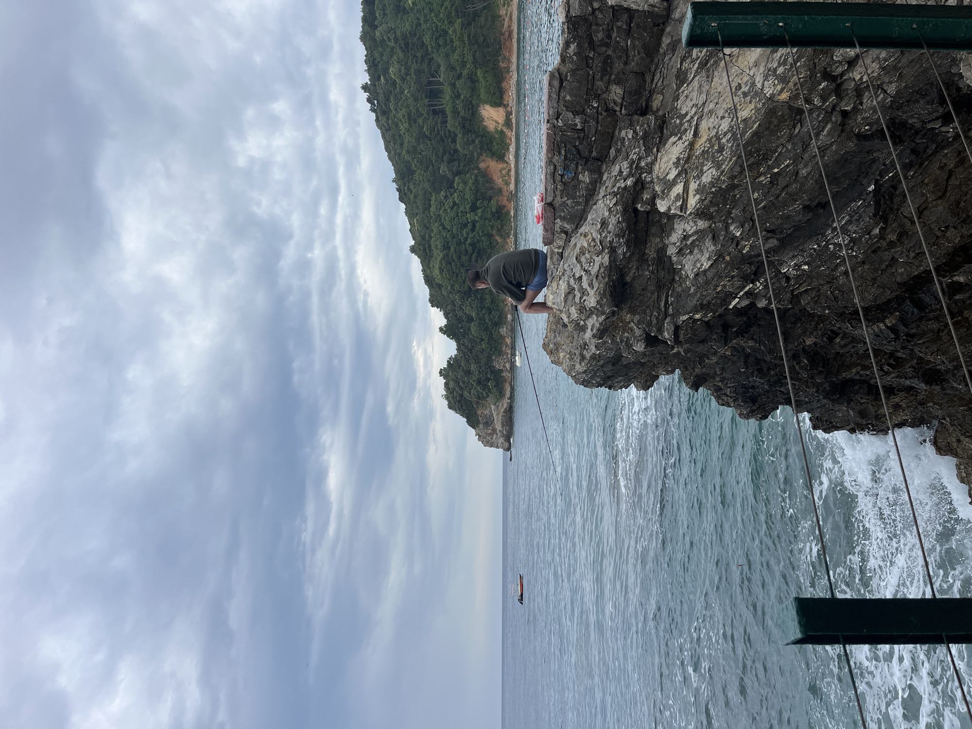 This image, captured in Budva, Montenegro, depicts an overcast coastal scene. The central subject is a man, dressed in a dark shirt and shorts, perched on a large, rugged rock formation at the edge of the sea. He appears to be fishing, holding a fishing rod that extends into the water below.
The setting is characterized by a rocky shoreline and calm, deep blue-green waters. To the left, a lush, tree-covered hill or island rises from the sea, presenting a natural green backdrop. In the mid-distance on the water, a small, slender orange boat or kayak is visible, along with several buoys scattered near the distant landmass.
On the right side of the frame, dark, textured rock cliffs dominate, with waves breaking against their base. A dark green metal beam or railing is visible above these rocks, from which several thin metal cables or wires hang downwards. The sky above is overcast, filled with soft grey and white clouds, suggesting a cloudy daytime. The scene evokes a sense of peaceful contemplation, centered around the solitary activity of fishing in a serene natural environment. No visible text is present in the image.