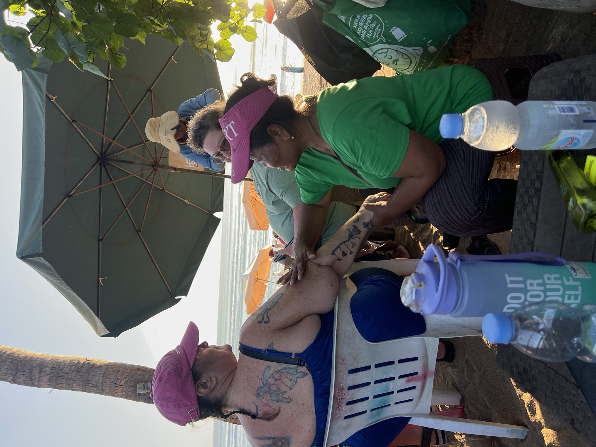 This is a medium-shot, eye-level outdoor photograph taken in Kuta, Indonesia, likely on the beach given the presence of sand, palm trees, and the ocean in the background. The image features a group of people under a large green umbrella. In the foreground, a woman with dark hair and wearing a green t-shirt and a pink visor is getting her arm tattooed by another person. The tattooed individual, a woman wearing a blue swimsuit and a pink baseball cap, is seated in a white plastic chair, facing away from the camera. Her back and arm are covered in tattoos, including a large sea turtle on her back. In the background, a person wearing a wide-brimmed hat and a denim jacket is visible standing under the umbrella. The scene is illuminated by the warm glow of sunset, casting long shadows and highlighting the textured surfaces. There are several water bottles visible on a table in the foreground. The overall atmosphere appears relaxed and casual.